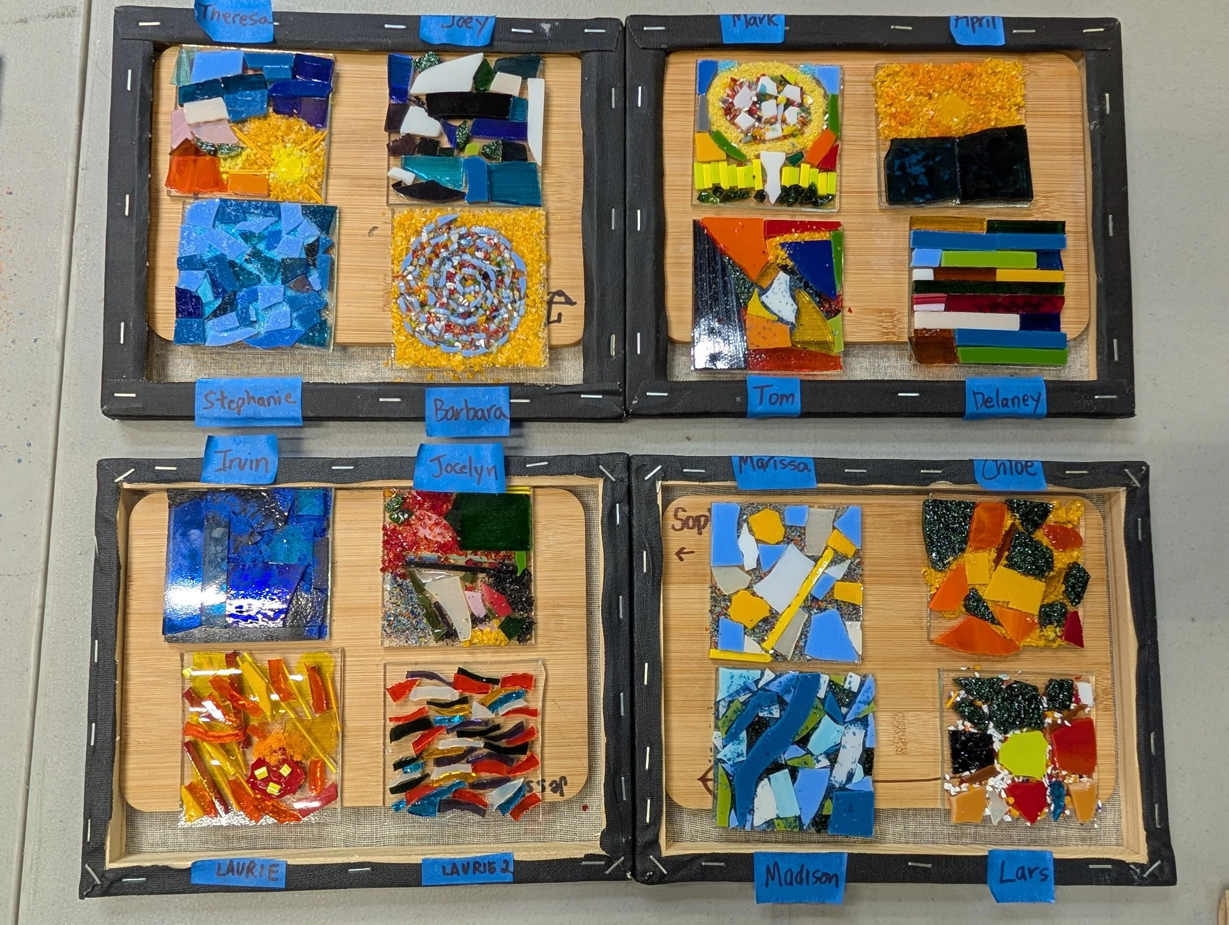 Four rectangular display boxes contain colorful mosaic glass art pieces, each labeled with a child's name. The top left box has mosaics labeled 'Theresa', 'Joey', and 'Mark'. Teambuilding event for groups.