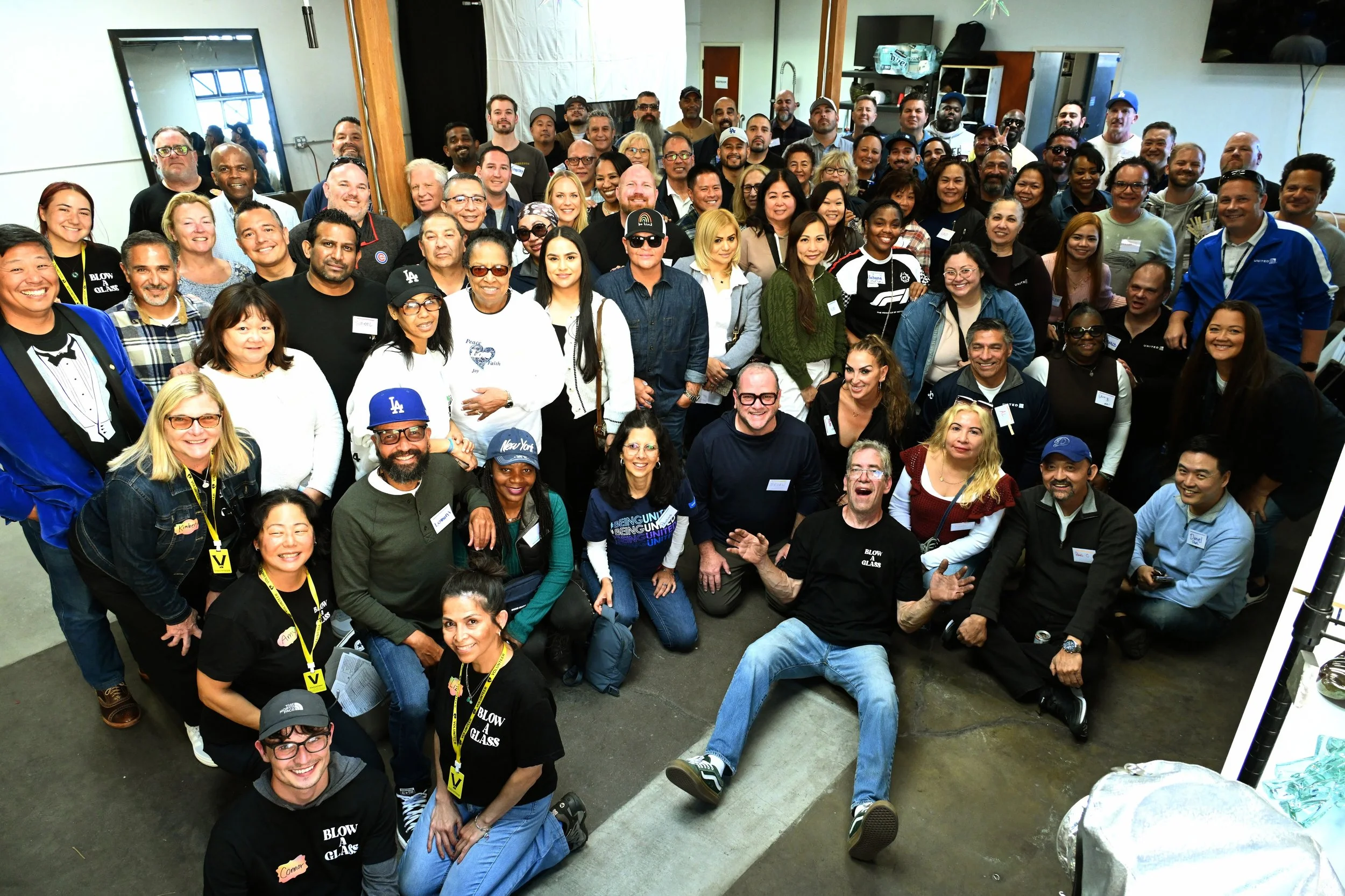 A large group of diverse people gathered together indoors for a group photo, smiling and facing the camera, with some wearing name tags and casual attire.