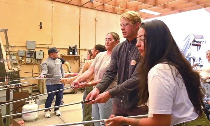 People standing in a line, holding long metal rods, in a workshop or factory setting for a hands-on activity. Teambuilding event for groups.