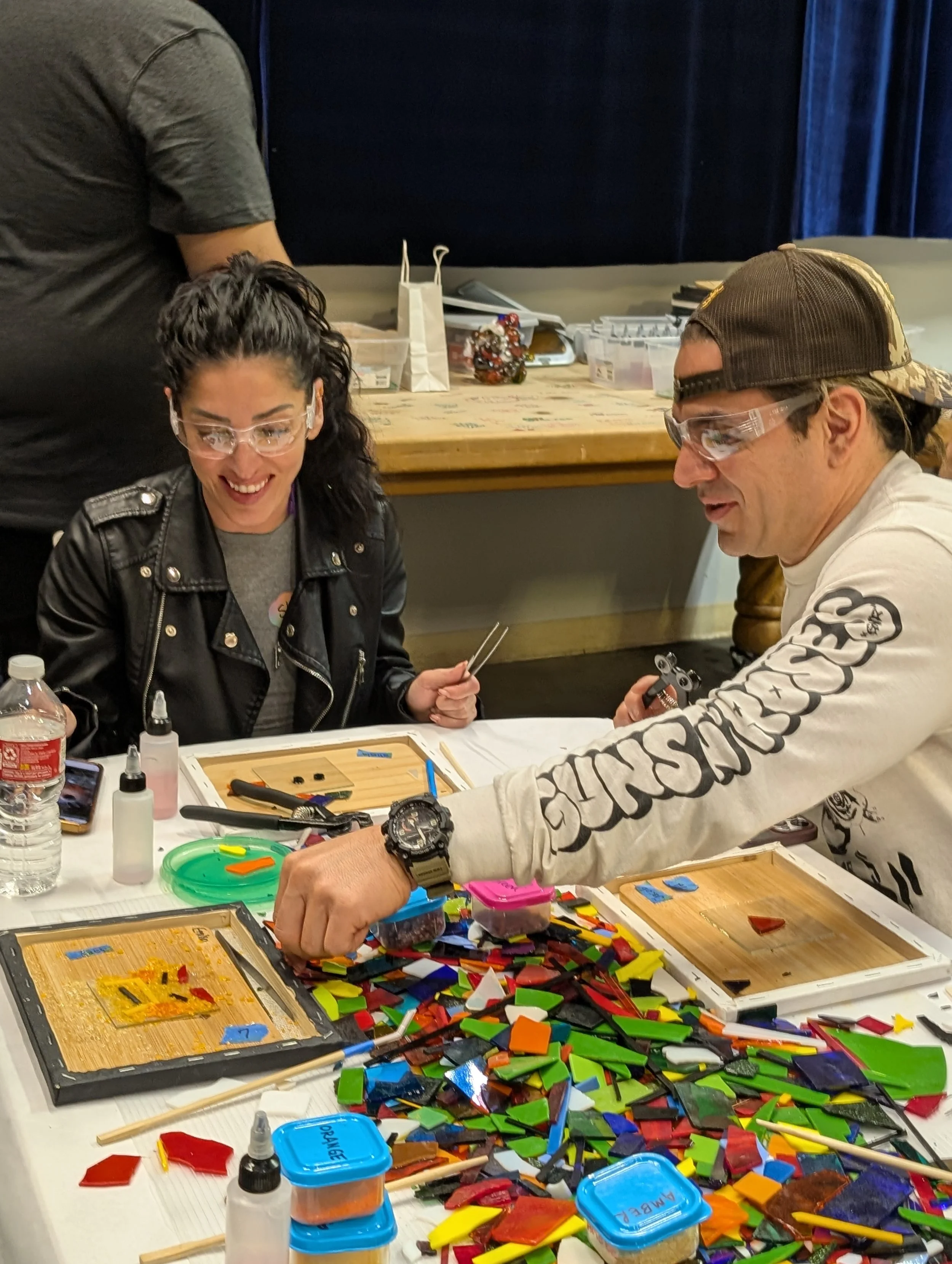 Two people at a table working on stained glass projects, surrounded by colorful glass pieces and tools.