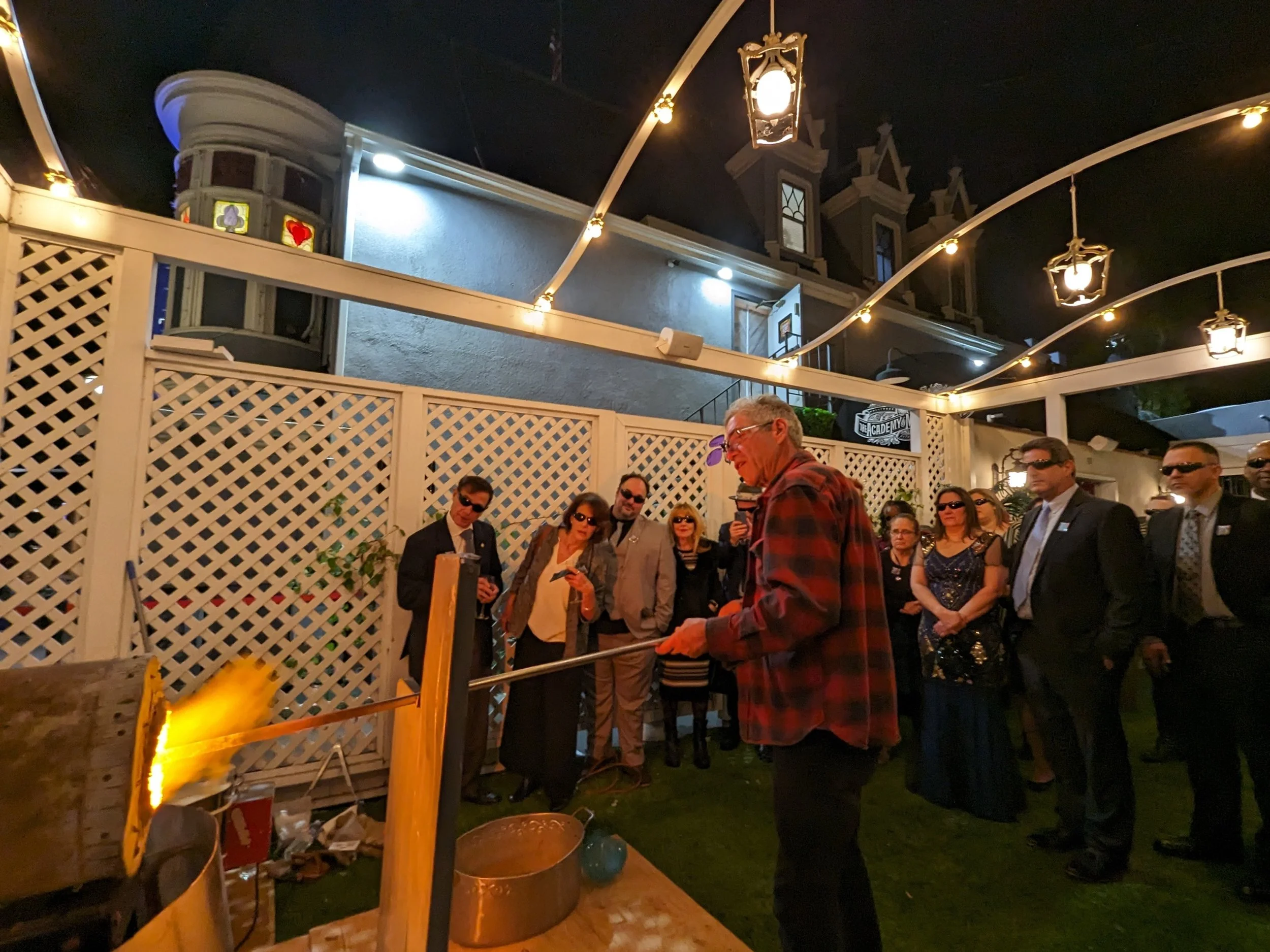 A man wearing a red and black plaid shirt is demonstrating a science experiment involving fire, while a group of people in formal attire and sunglasses watches at night outside. Corporate team building.