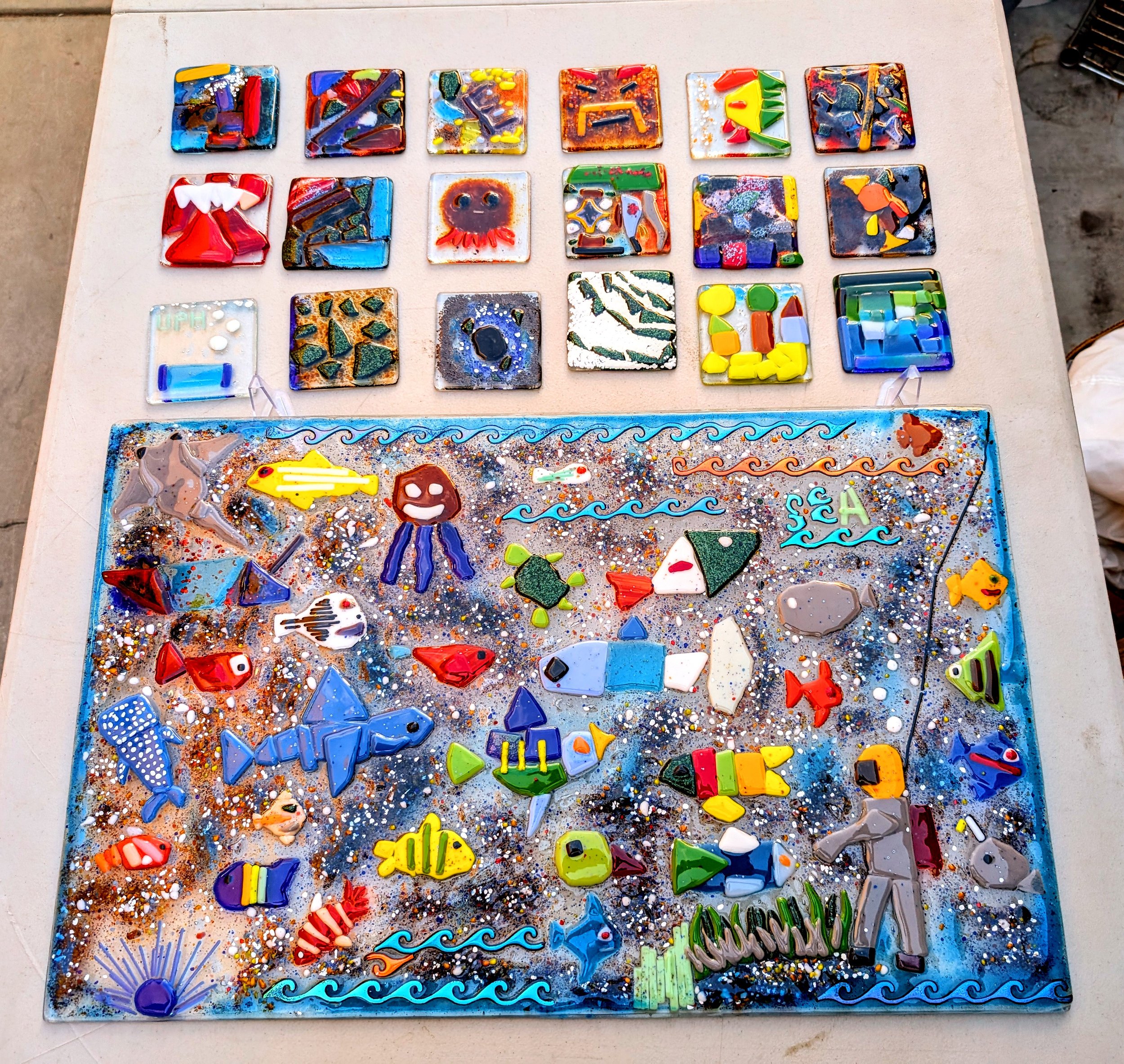 Colorful glass mosaic art pieces, including sea animals, marine life, and abstract designs, displayed on a table.
