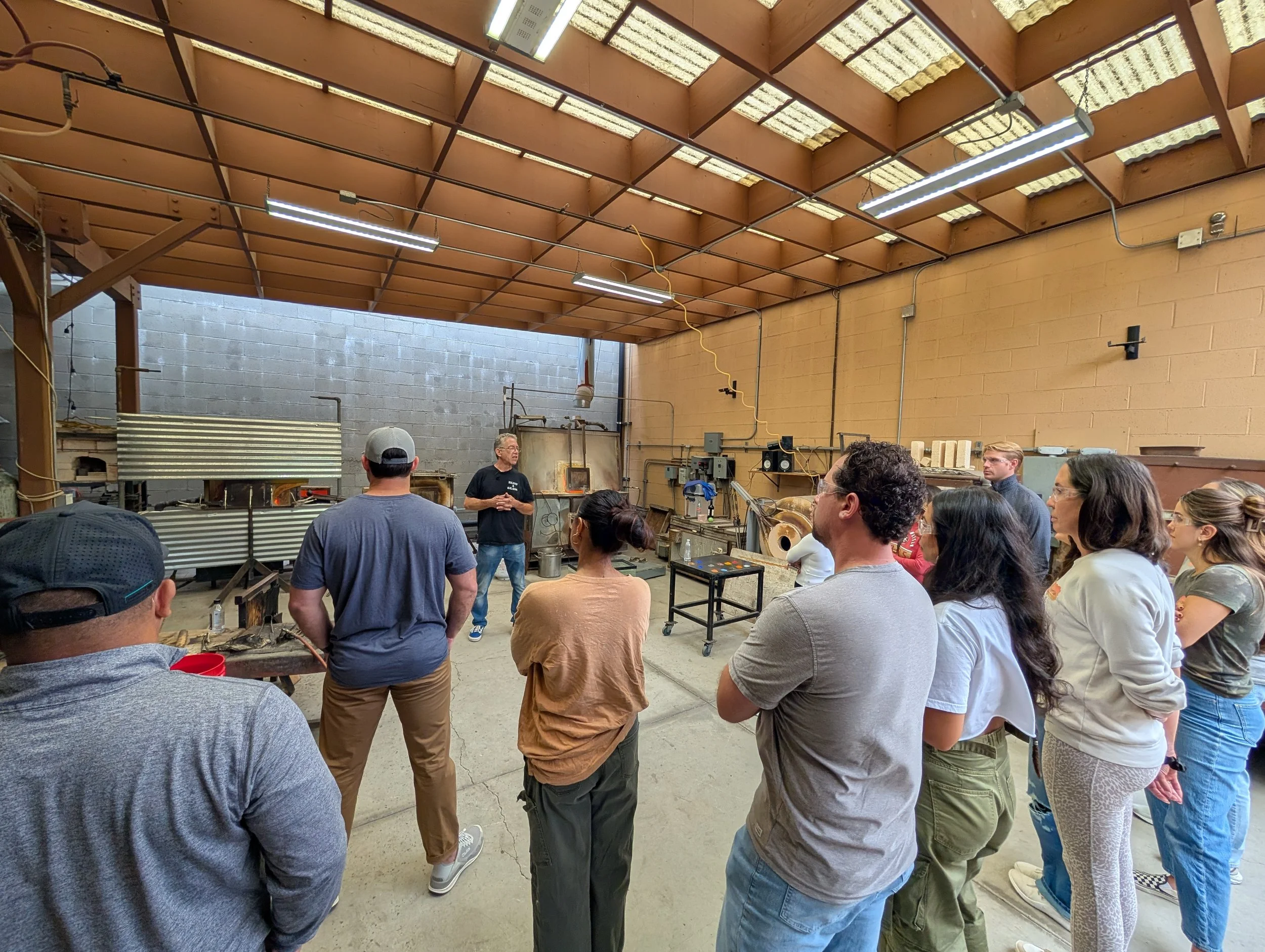 A group of people listening to a man giving a talk or presentation inside a workshop or industrial space, with industrial equipment and tools in the background. Teambuilding for groups and companies