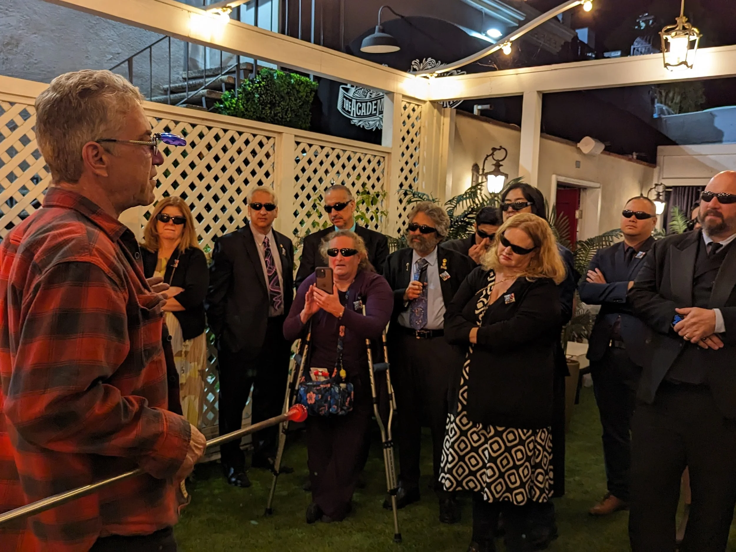 A man in a red plaid shirt gives a speech to a group of people wearing dark sunglasses at an outdoor gathering at night, with string lights and decorative plants in the background.