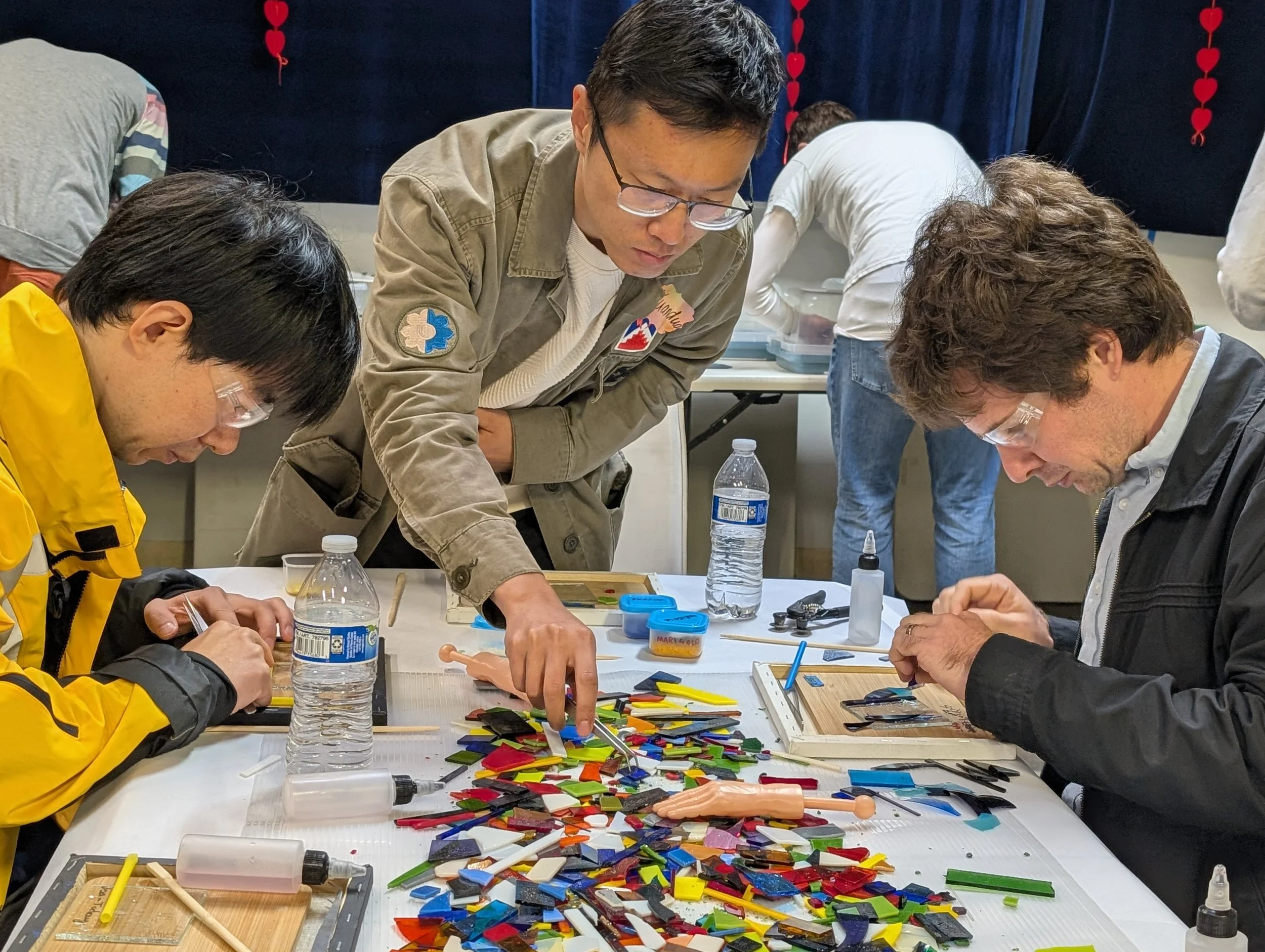 Three men working intently on a arts and crafts project involving colorful glass pieces, with tools and bottles of glue on the table.