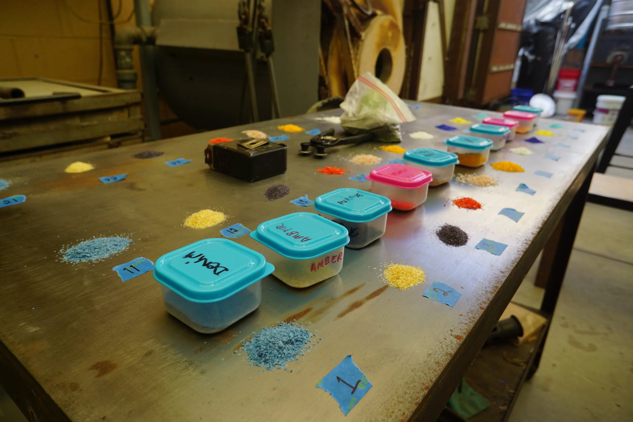 A wooden table with labeled containers of colored sand, each placed with corresponding colored paper labels for organizing or sorting, in a workshop or craft room setting.