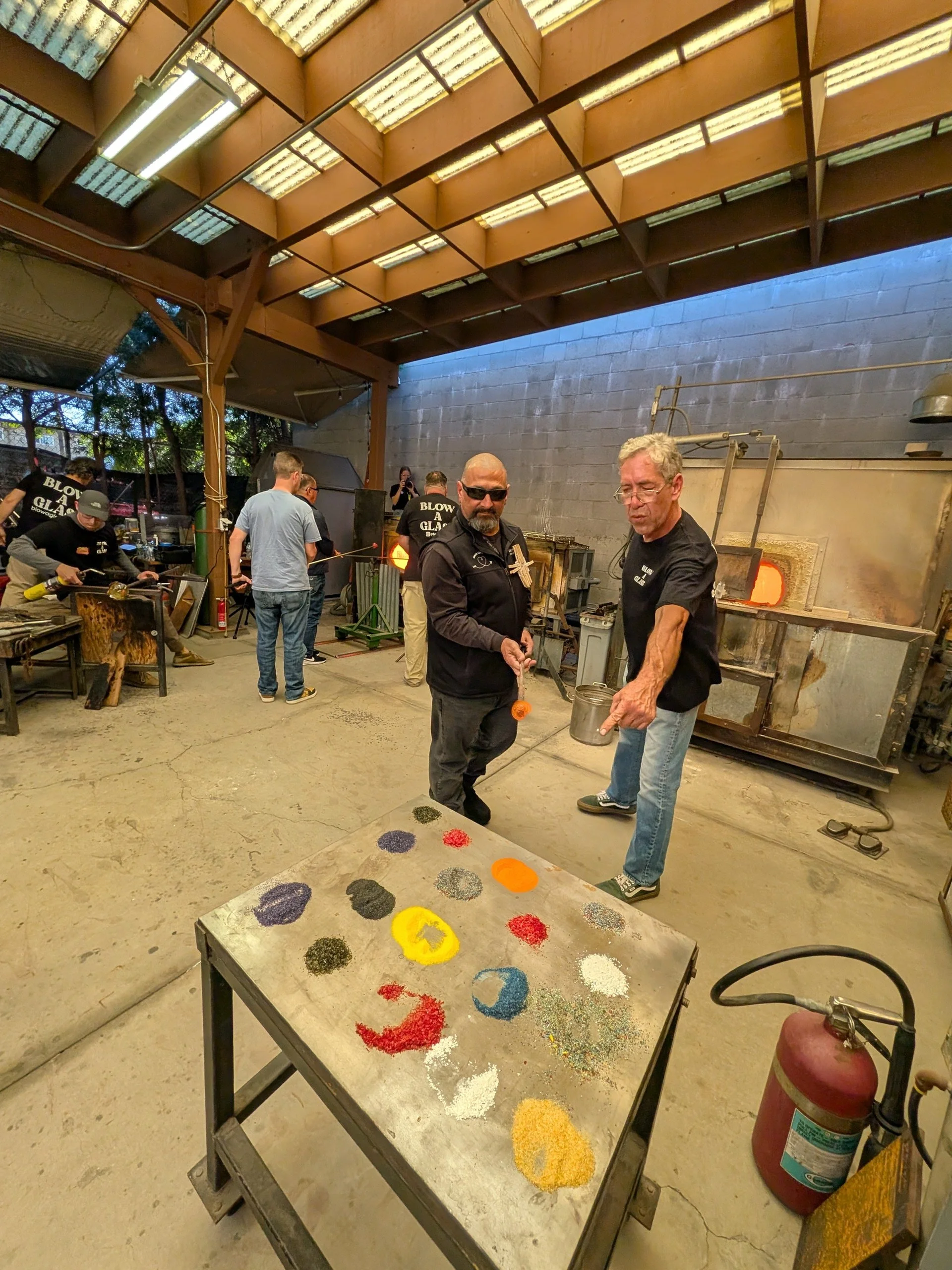 People in a glassblowing workshop, working with glowing glass in a furnace. A table with colorful glass powders in the foreground, and safety equipment including a fire extinguisher visible. Teambuilding event for groups.