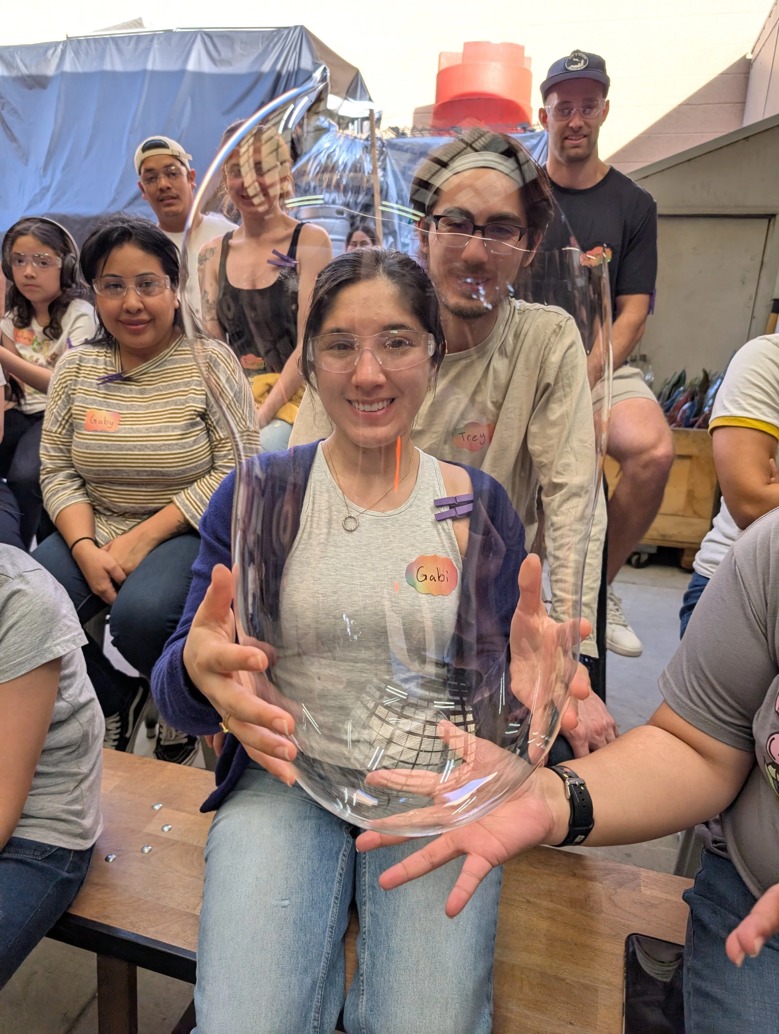 Group of people, some with glasses and name tags, gathered indoors, with one person holding a large, clear glass bowl or vessel. The central woman is smiling, and others are seated or standing around her, some behind her and some to her sides.