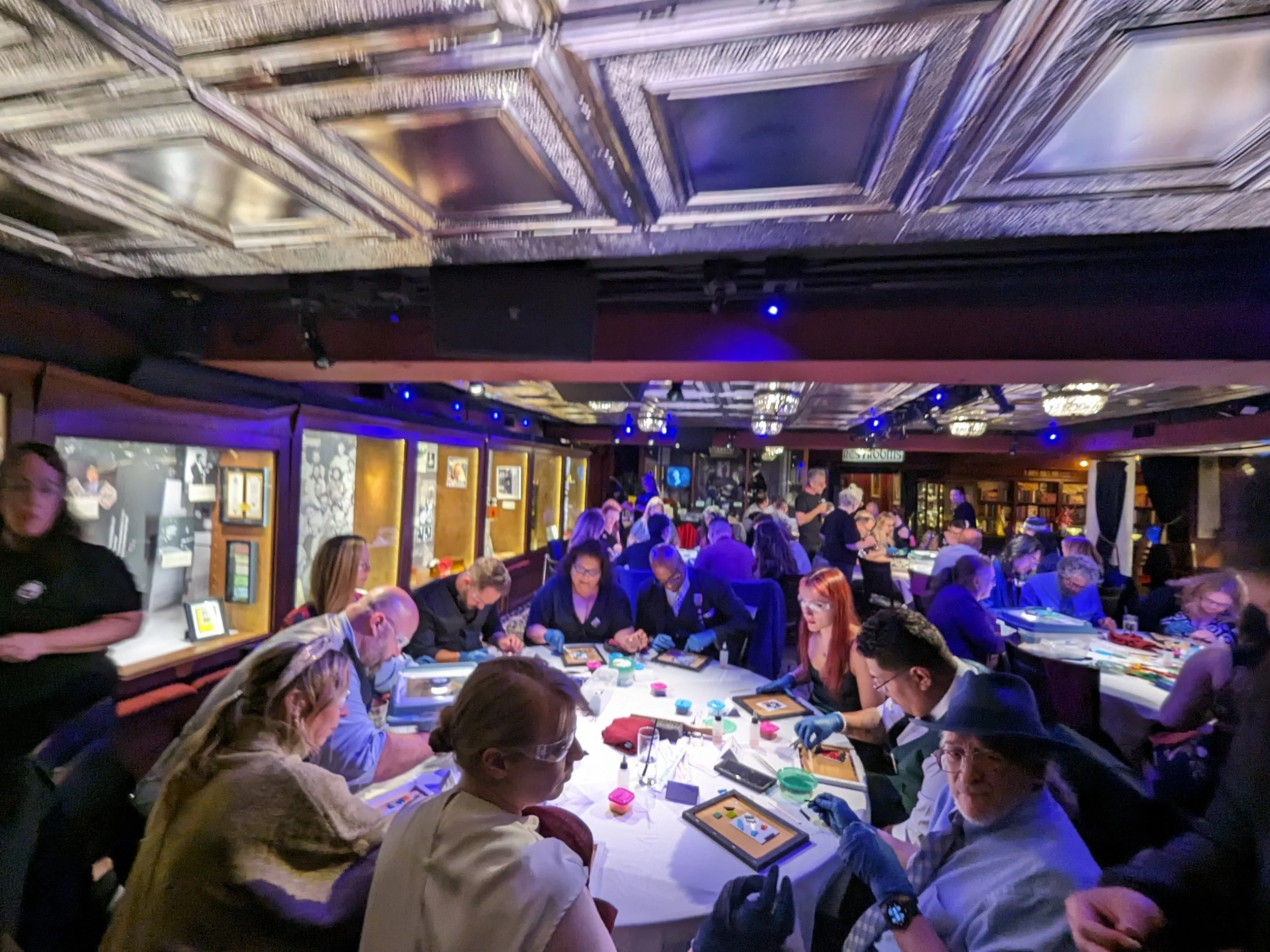 People playing a card game at multiple tables in a lively indoor venue with ornate ceiling and framed artwork on the walls.