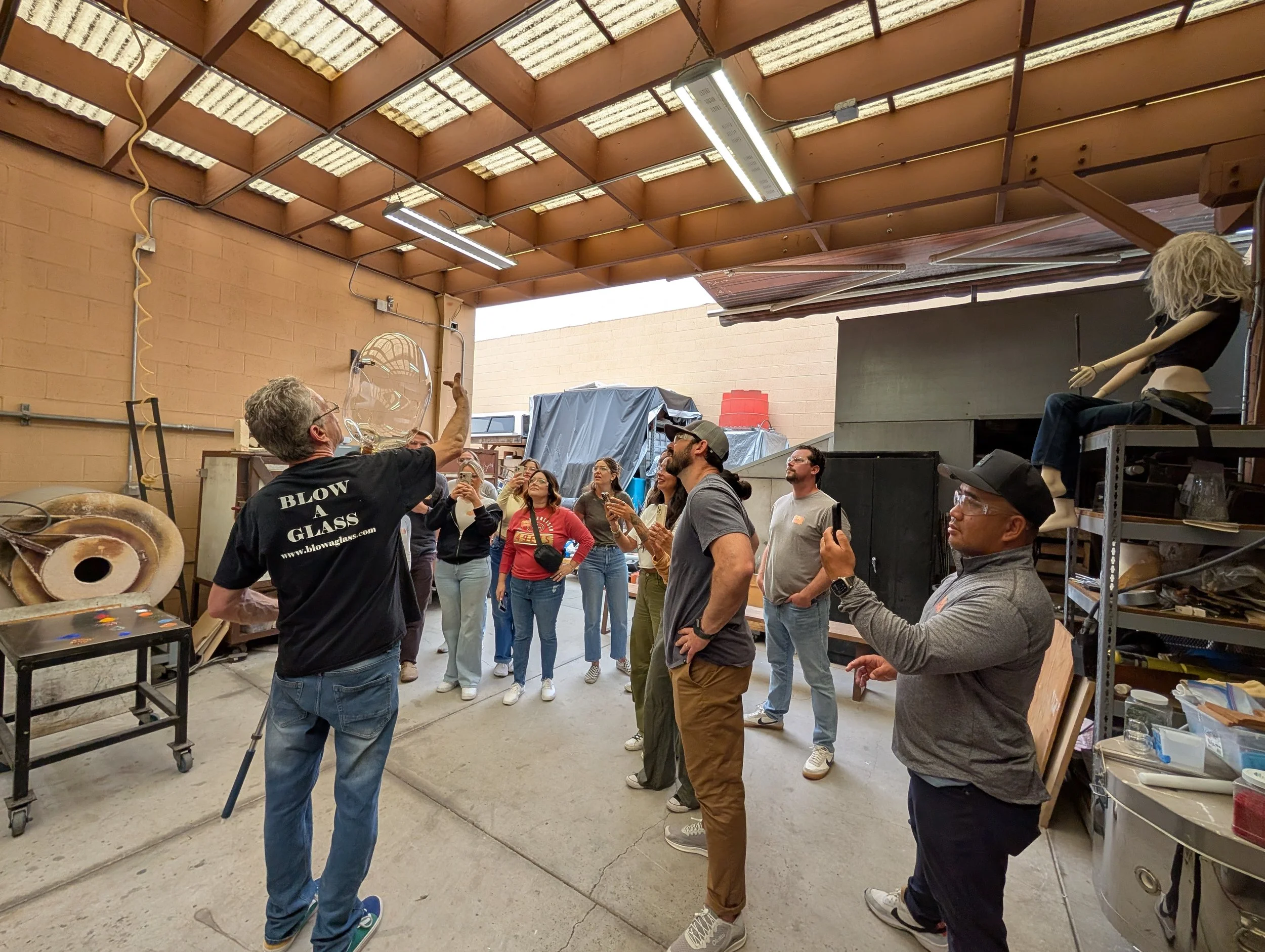 A man demonstrating glass blowing techniques to a group of people in a workshop with shelves and art supplies. Teambuilding event for groups.