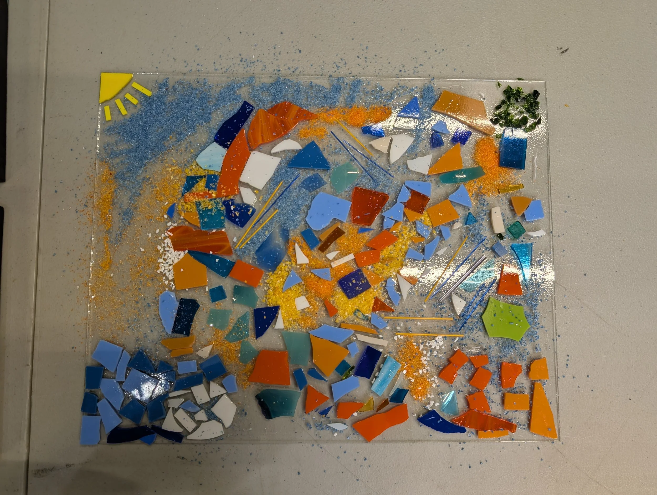 Colorful mosaic glass pieces on a transparent surface, with small details like a yellow sun shape in the top left corner and a green shape in the bottom right corner.