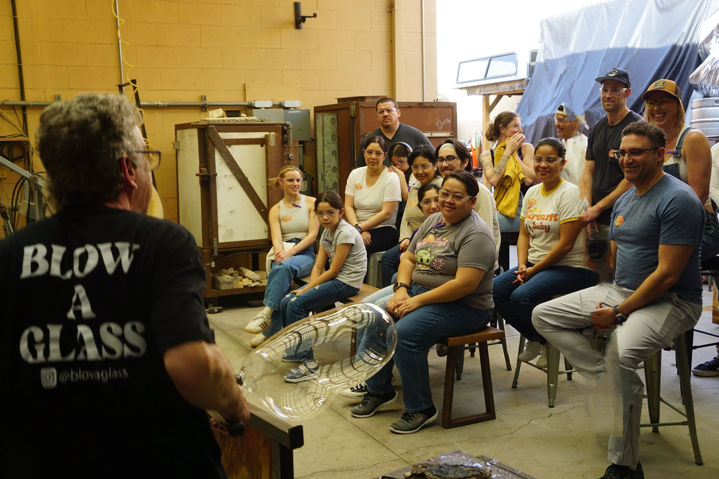 A glassblowing demonstration with an artist in a black shirt and an audience seated in a workshop space, watching a large, clear glass piece being formed.