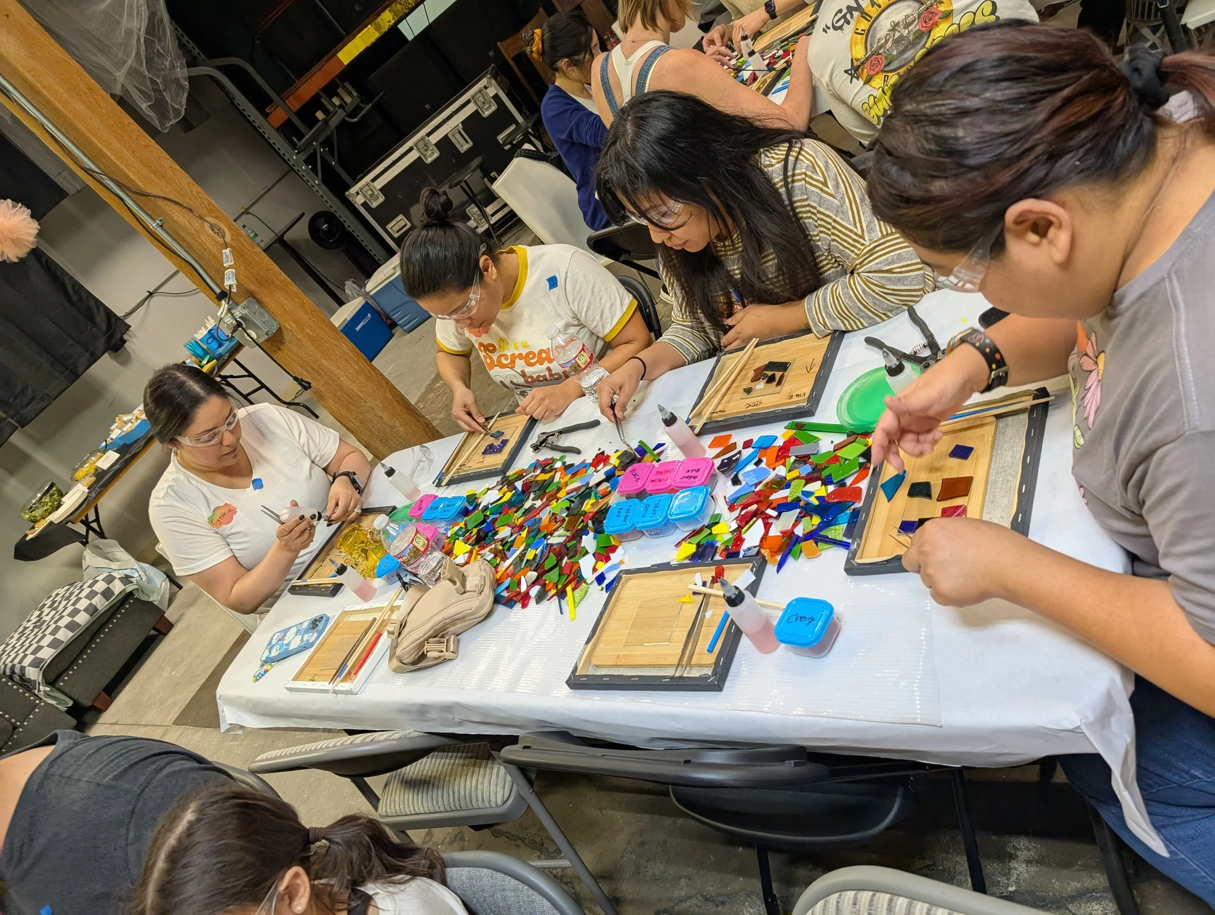 People sitting around a table working on glass mosaic art projects, using colorful glass pieces and tools. Teambuilding for groups and companies