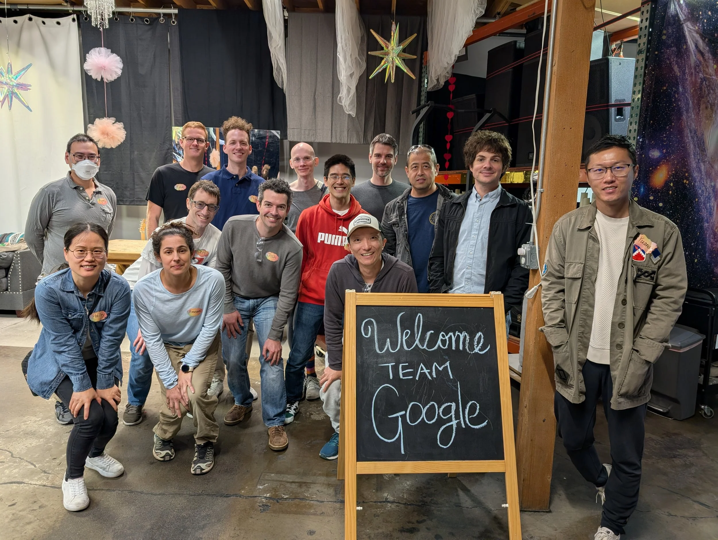 Group of people posing for a photo behind a chalkboard sign that reads "Welcome Team Google" in an indoor setting. Corporate Teambuilding event.