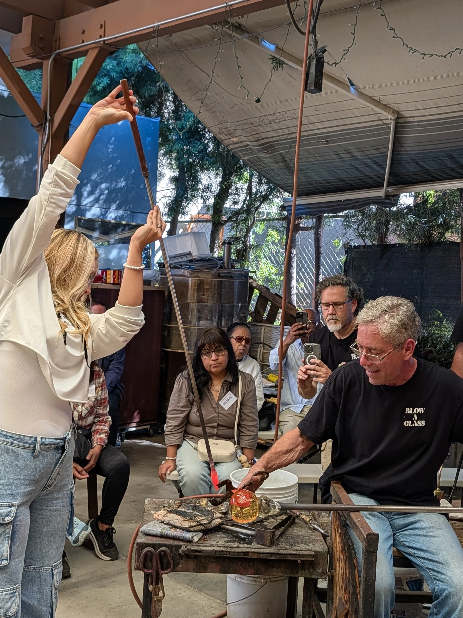 Glassblowing demonstration with a man shaping molten glass while others watch and record in a workshop. Corporate Team building event.
