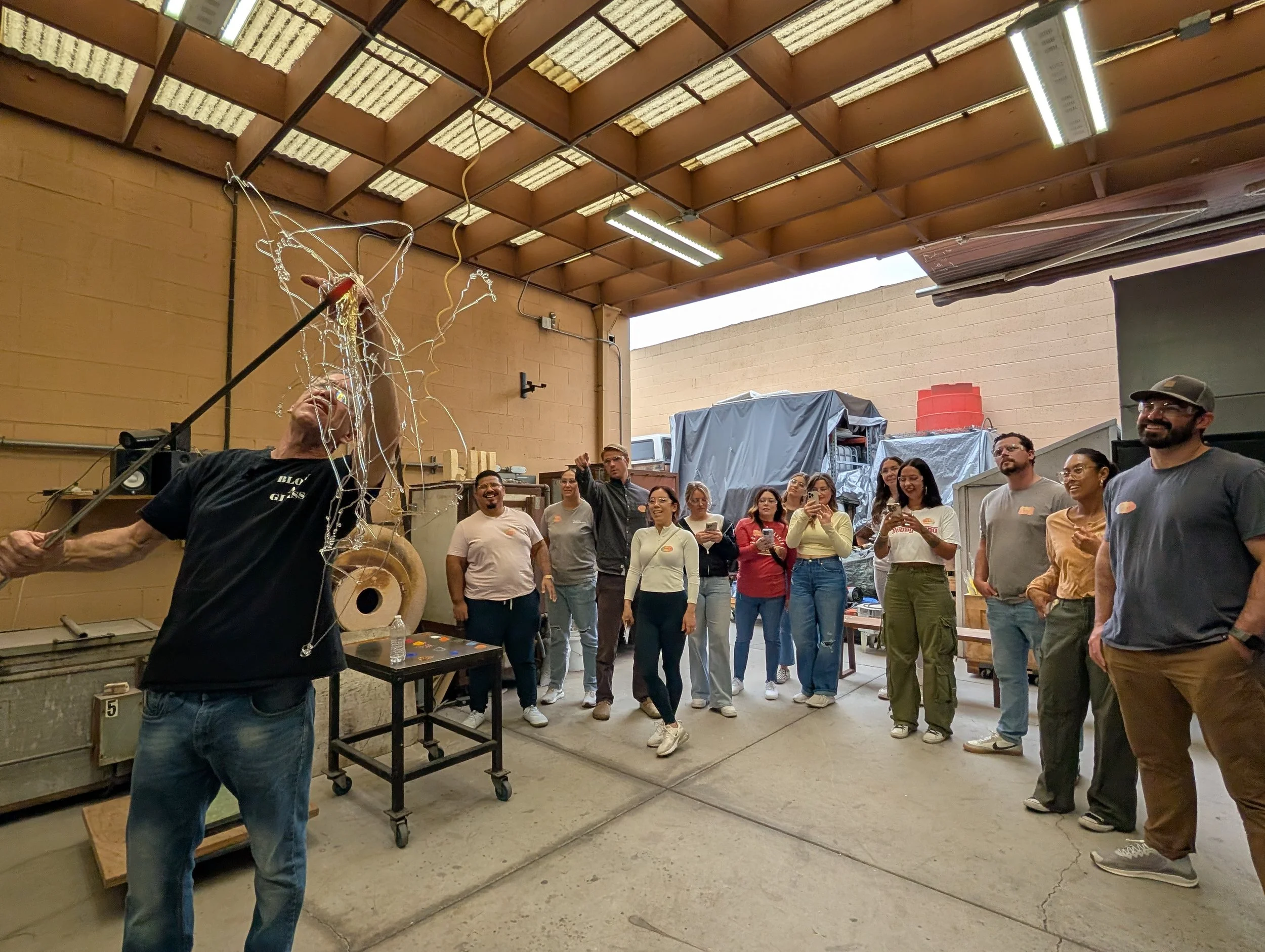 A man is holding a pole with a tangled mass of wire and objects suspended in mid-air, possibly in a workshop or garage. A group of people, mostly smiling and watching, stand in a semi-circle behind him. Teambuilding event for groups.