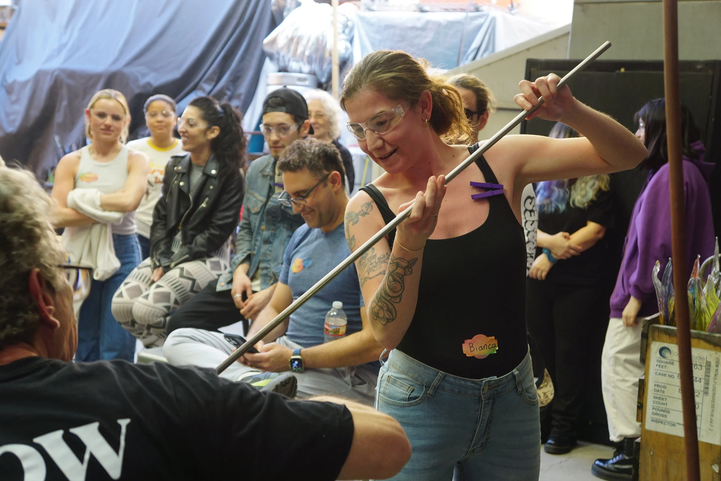 A woman with tattoos and glasses holding a long stick or pole, interacting with a person sitting down at an event or gathering with several onlookers in the background.