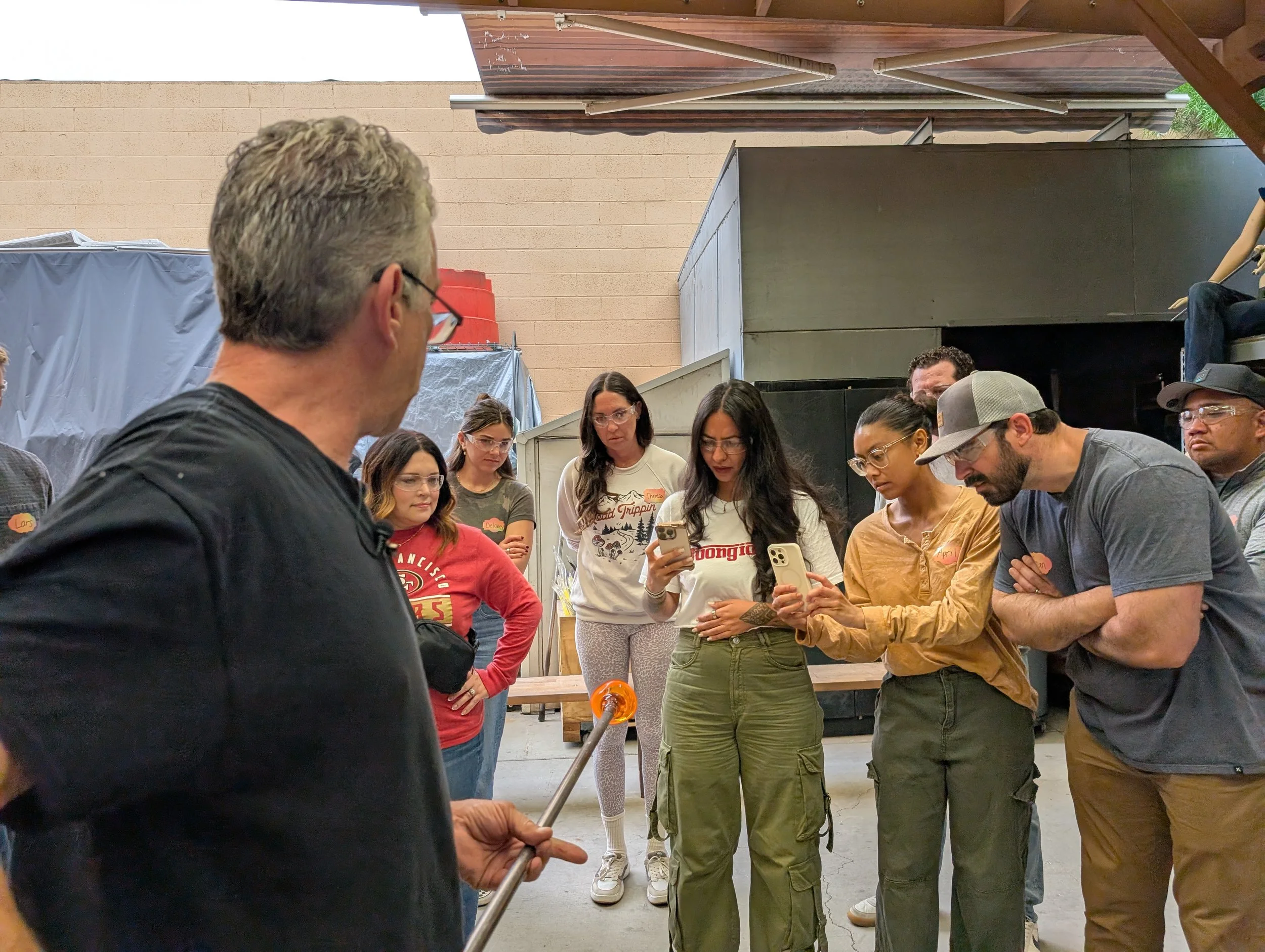 A group of people gathered around a man holding a halogen torch, possibly in a workshop or outdoor setting, with some people looking at their phones and others paying attention to the man.