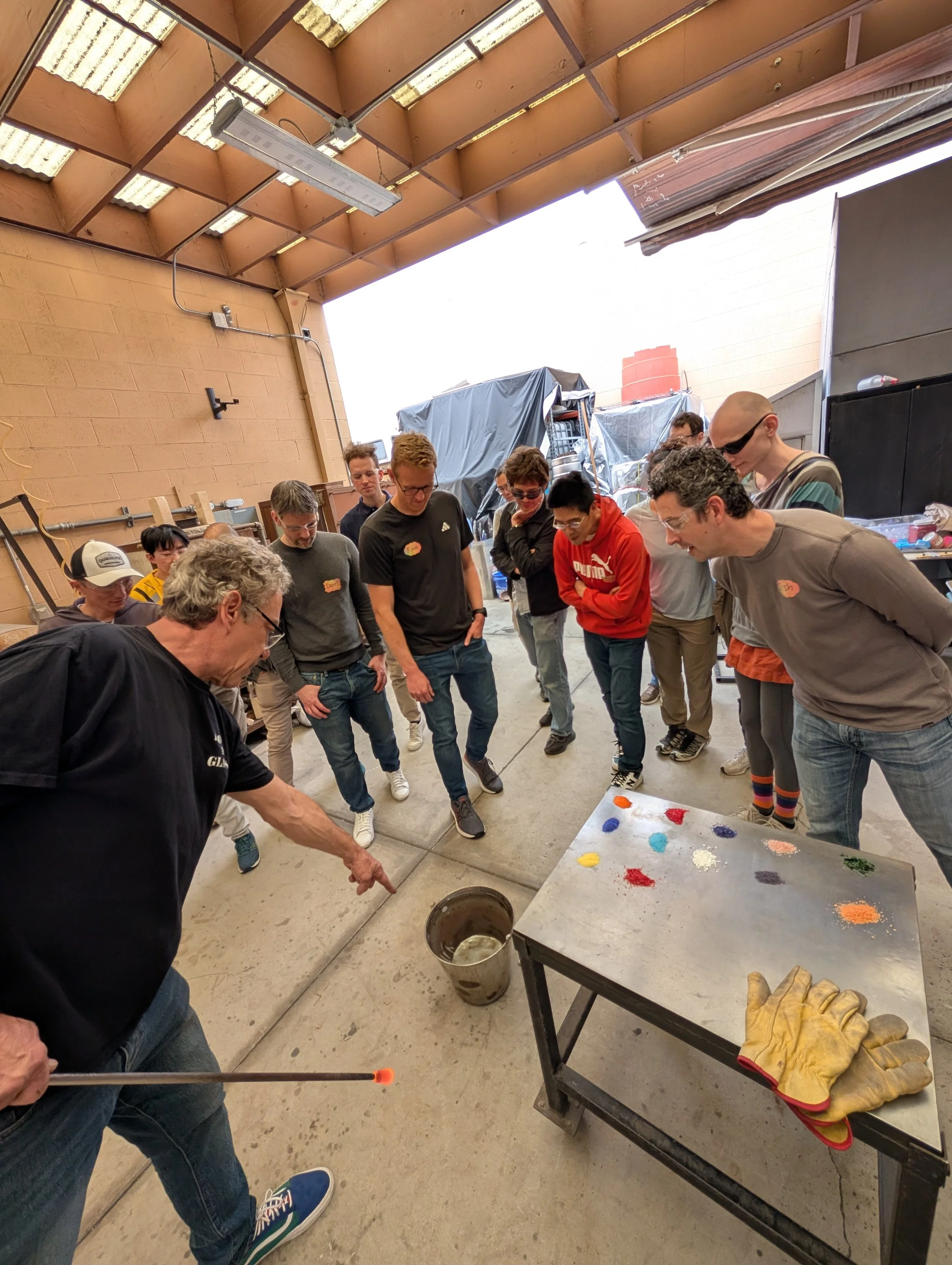Group of people gathered around a table outdoors in a garage or workshop, observing a demonstration by an older man pointing to an object on the ground. There are gloves and colorful powders on the table.