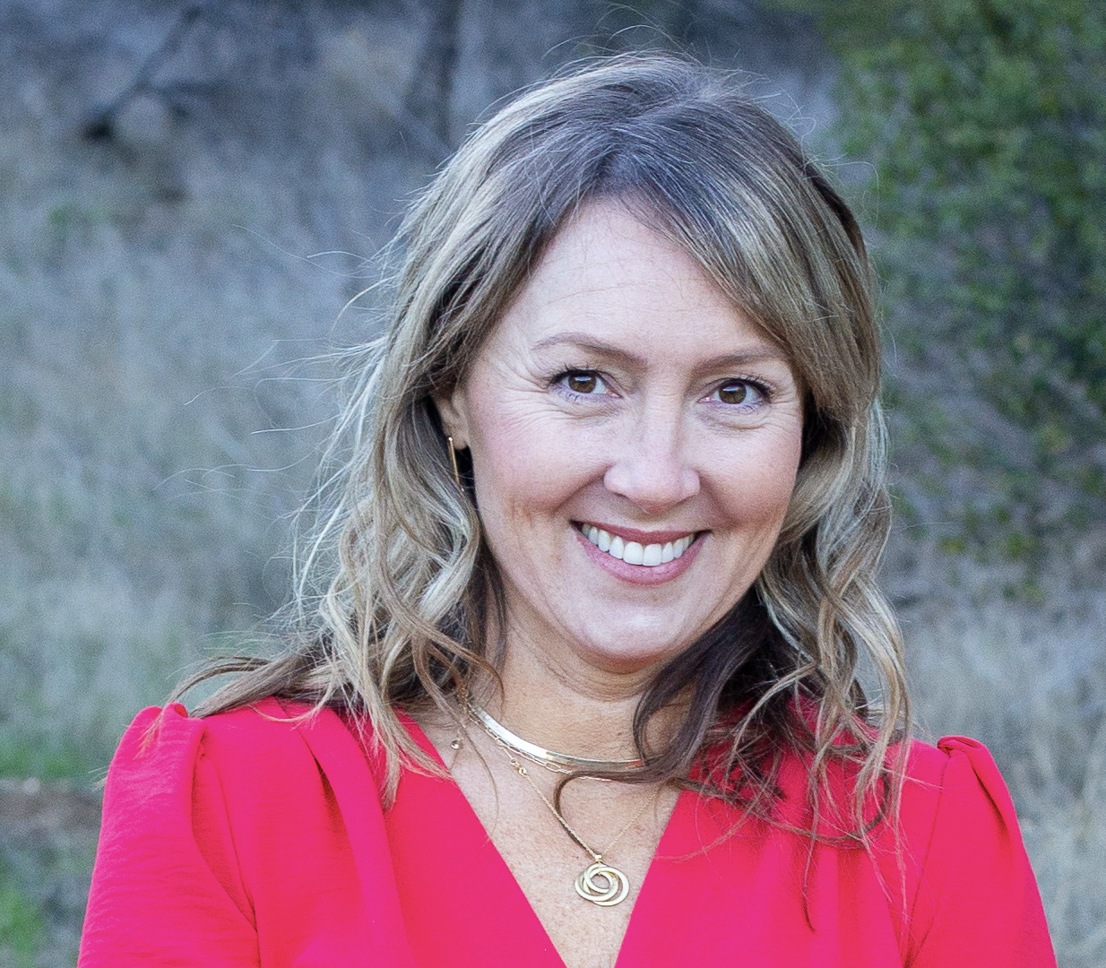A woman with shoulder-length light brown hair and light skin, smiling outdoors in front of a grassy background, wearing a red top and gold jewelry.
