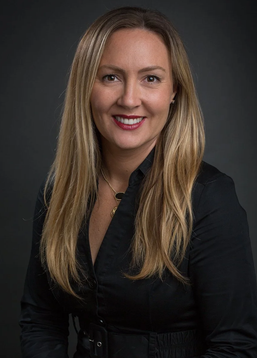 A woman with shoulder-length light brown hair and light skin, smiling outdoors in front of a grassy background, wearing a red top and gold jewelry.