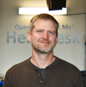 A man with short blond hair and facial hair, smiling in front of a sign that says "Hey, Desk."