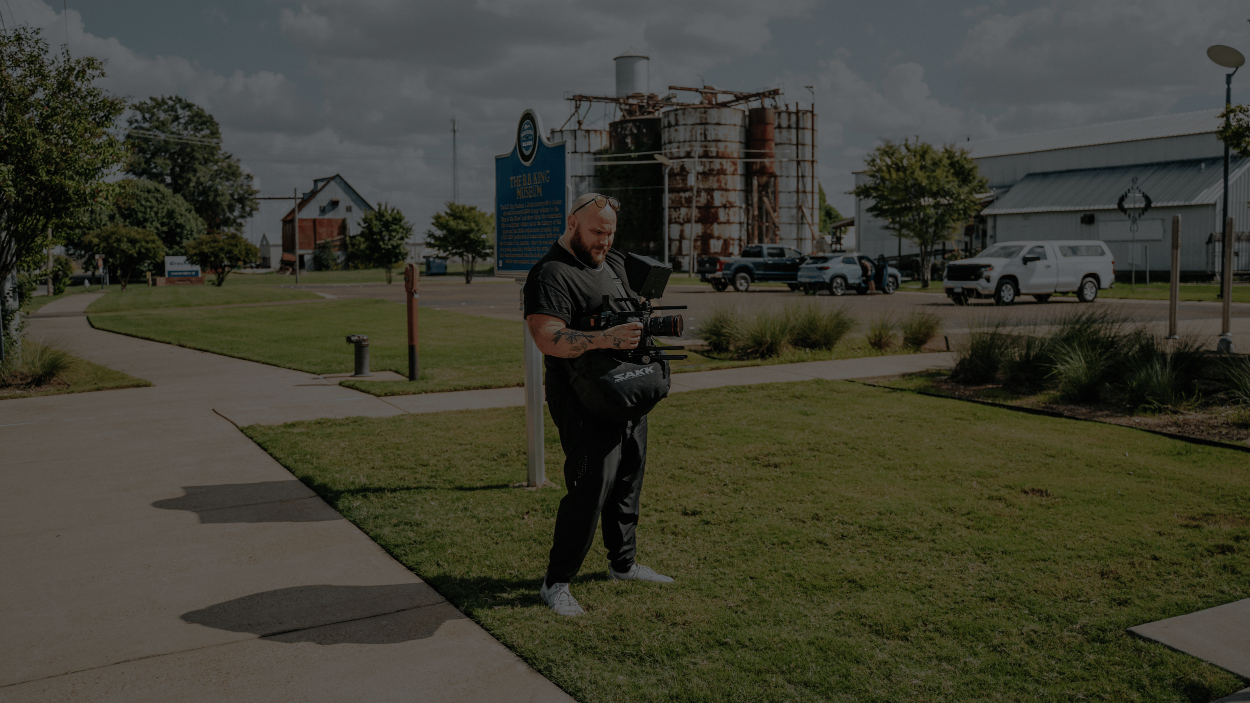 Smiling man with a beard and nose piercing, wearing a white short-sleeve button-up shirt, standing against a rocky background.
