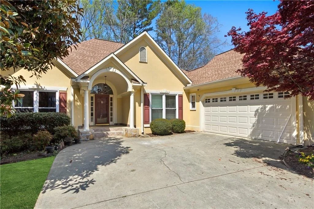 Yellow single-family house with red shutters, a garage, and a front porch with columns, surrounded by trees and bushes, on a sunny day.