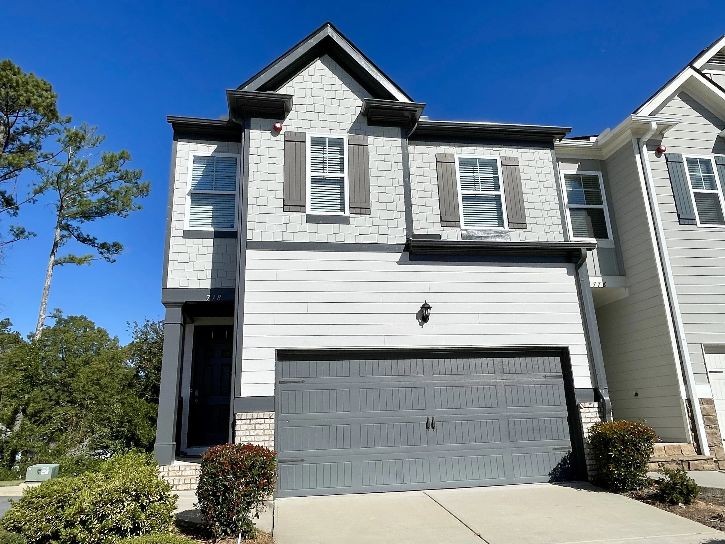 Front of a modern two-story house with gray exterior, black roof, and closed garage door, surrounded by shrubs and trees under a clear blue sky.
