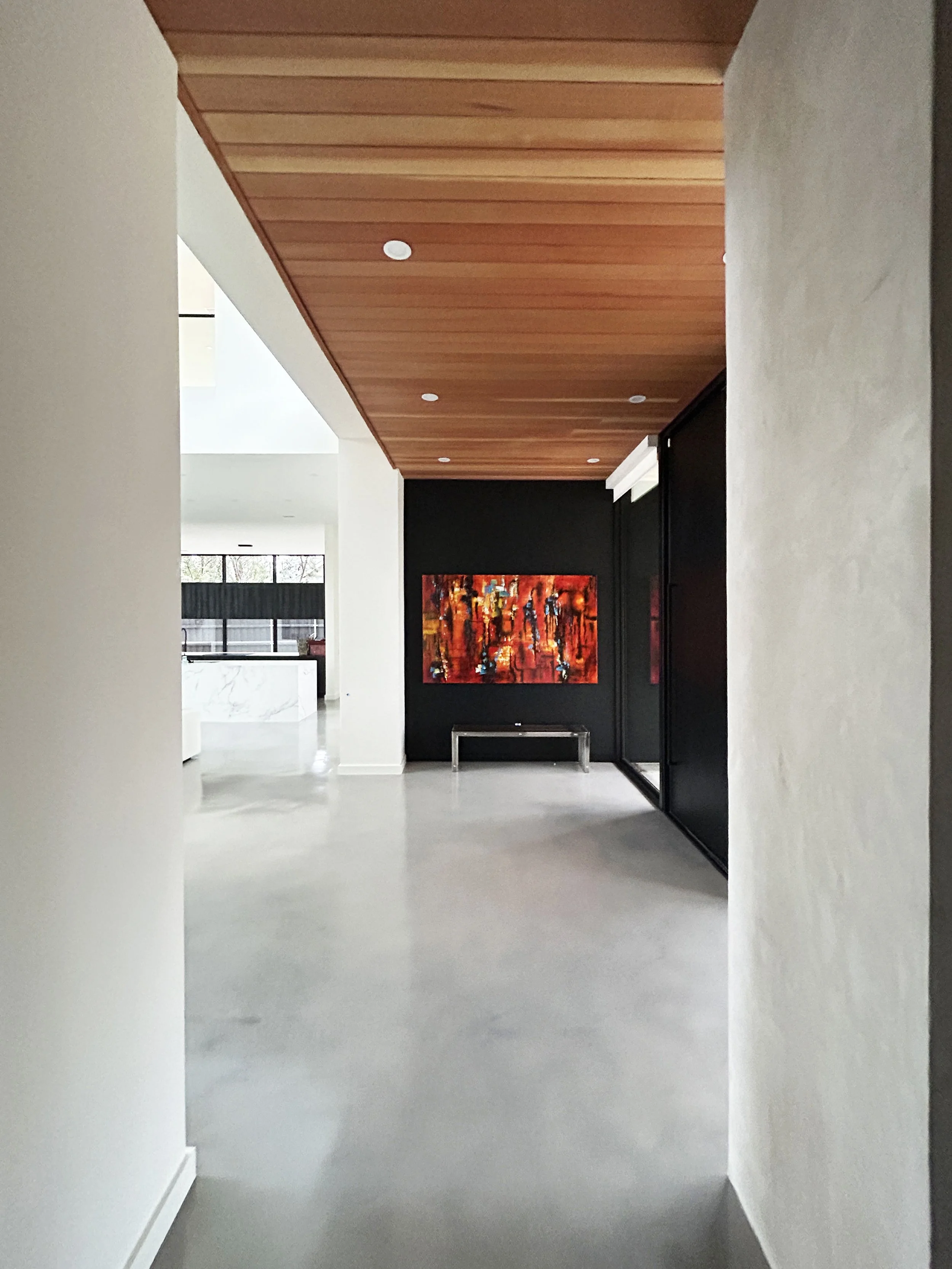 Luxury modern home interior featuring a minimalist entry hallway with warm wood-paneled ceilings, crisp white walls, polished concrete flooring, and dramatic black accent walls. A vibrant abstract red and orange artwork adds a bold focal point, compl