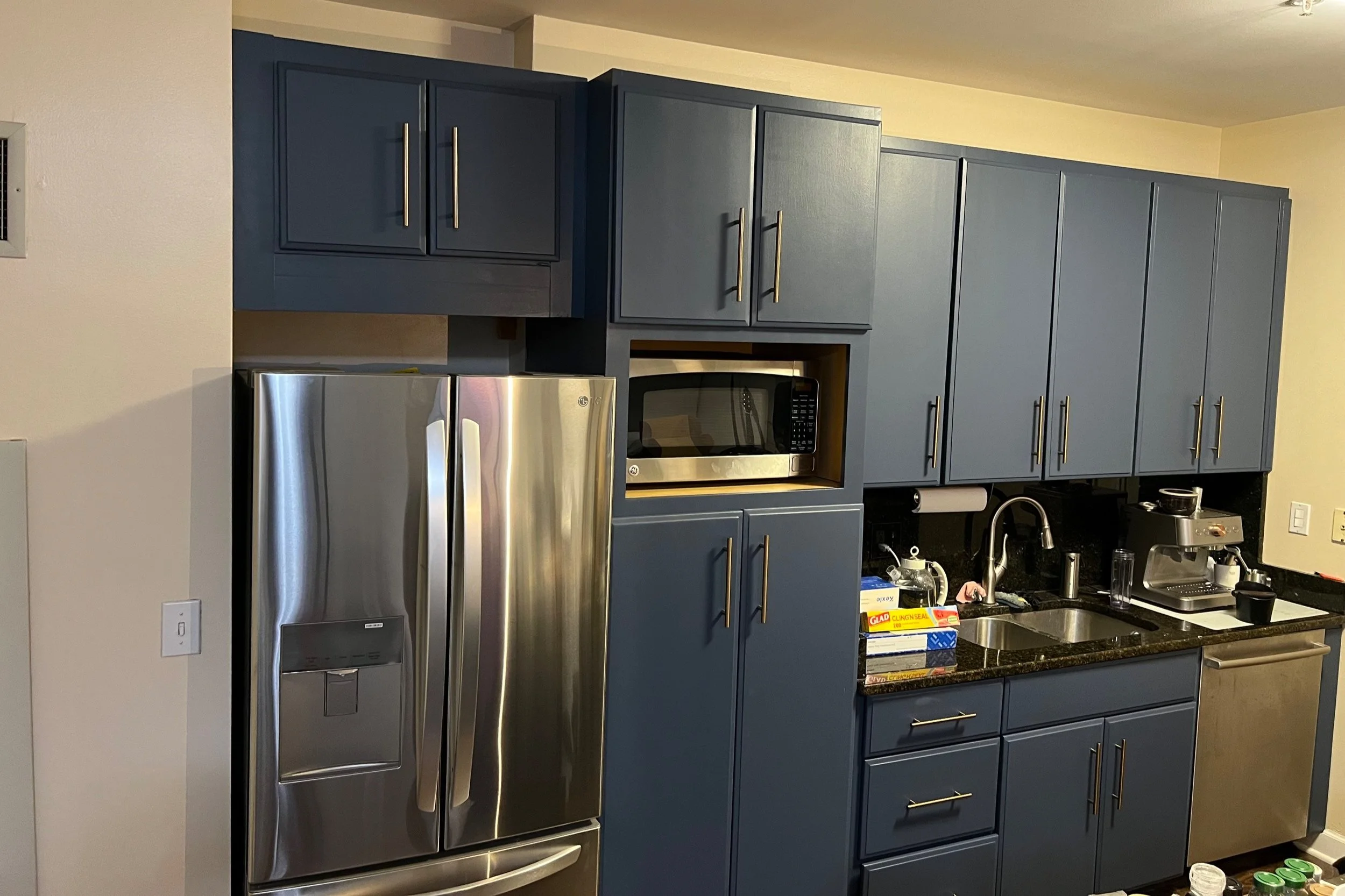 Kitchen with blue cabinets, stainless steel refrigerator, microwave, sink, coffee maker, and boxes of Glad Cling Wrap on black granite countertop.