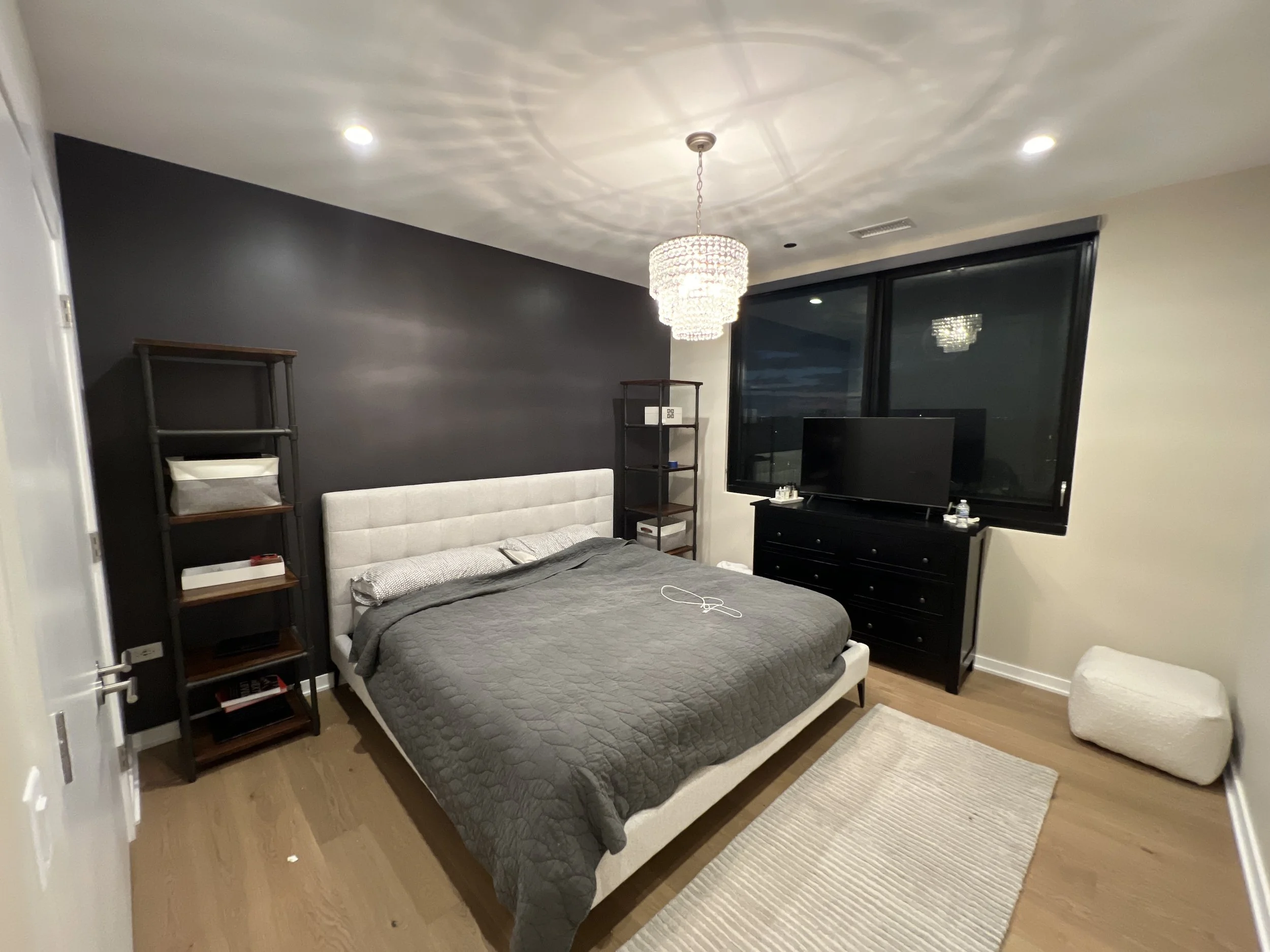 Modern bedroom with a white upholstered bed, dark gray quilt, black shelving units, black dresser with a TV, large window, and a white textured ottoman.
