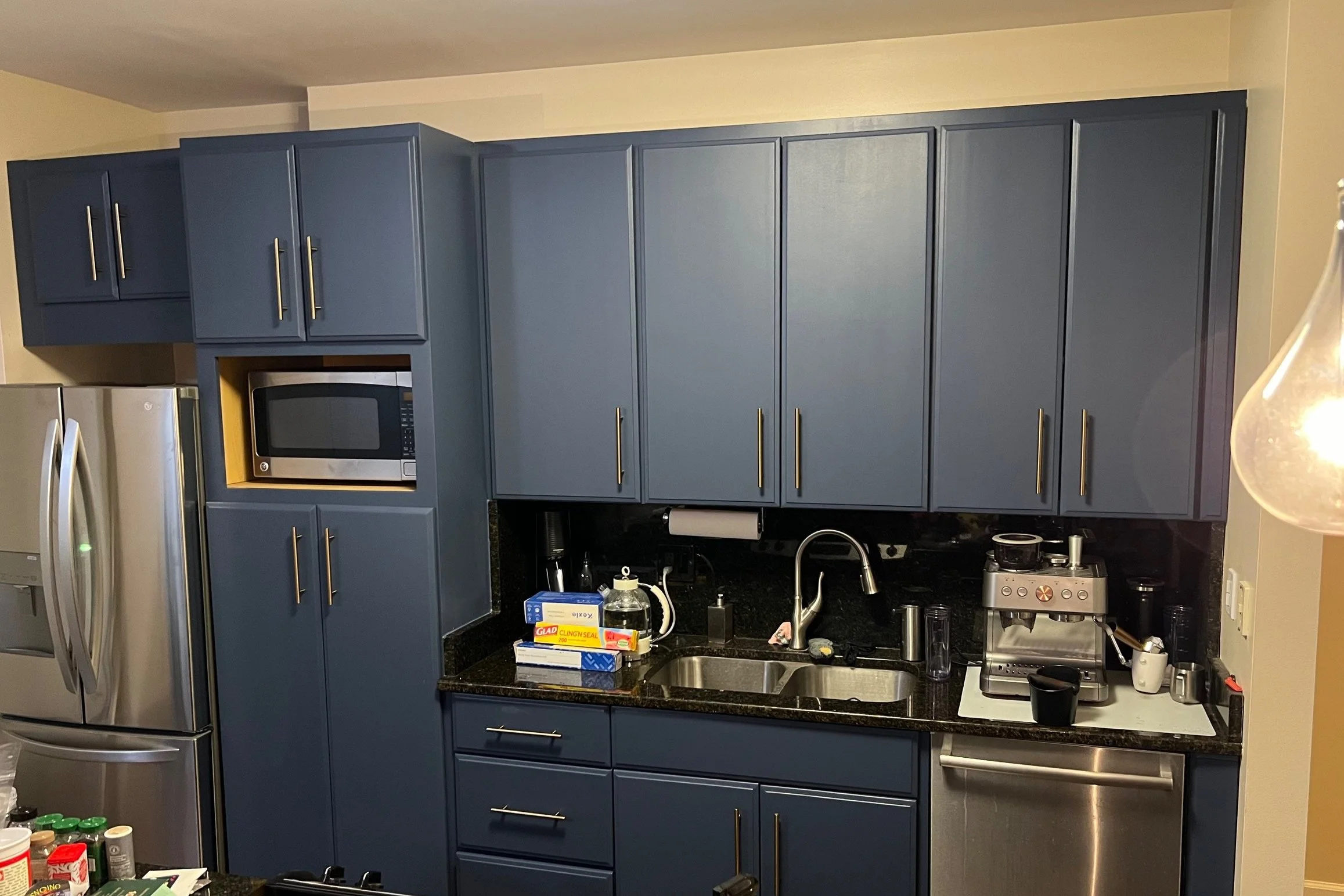 Kitchen with blue cabinets, black granite countertop, stainless steel appliances including a refrigerator, microwave, and dishwasher. Items on the counter include a coffee machine, papers, and various kitchen supplies.