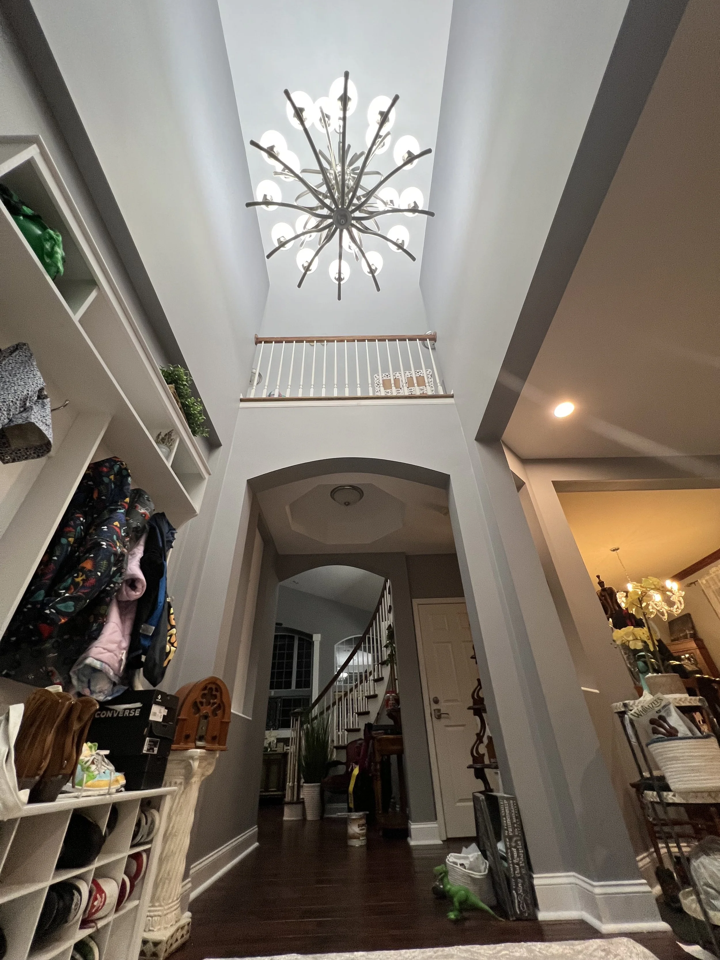 View of a two-story foyer with a modern chandelier, a staircase with white railings, and an interior door. Shelving with shoes and backpacks on the left, and a dining area with a chandelier on the right.