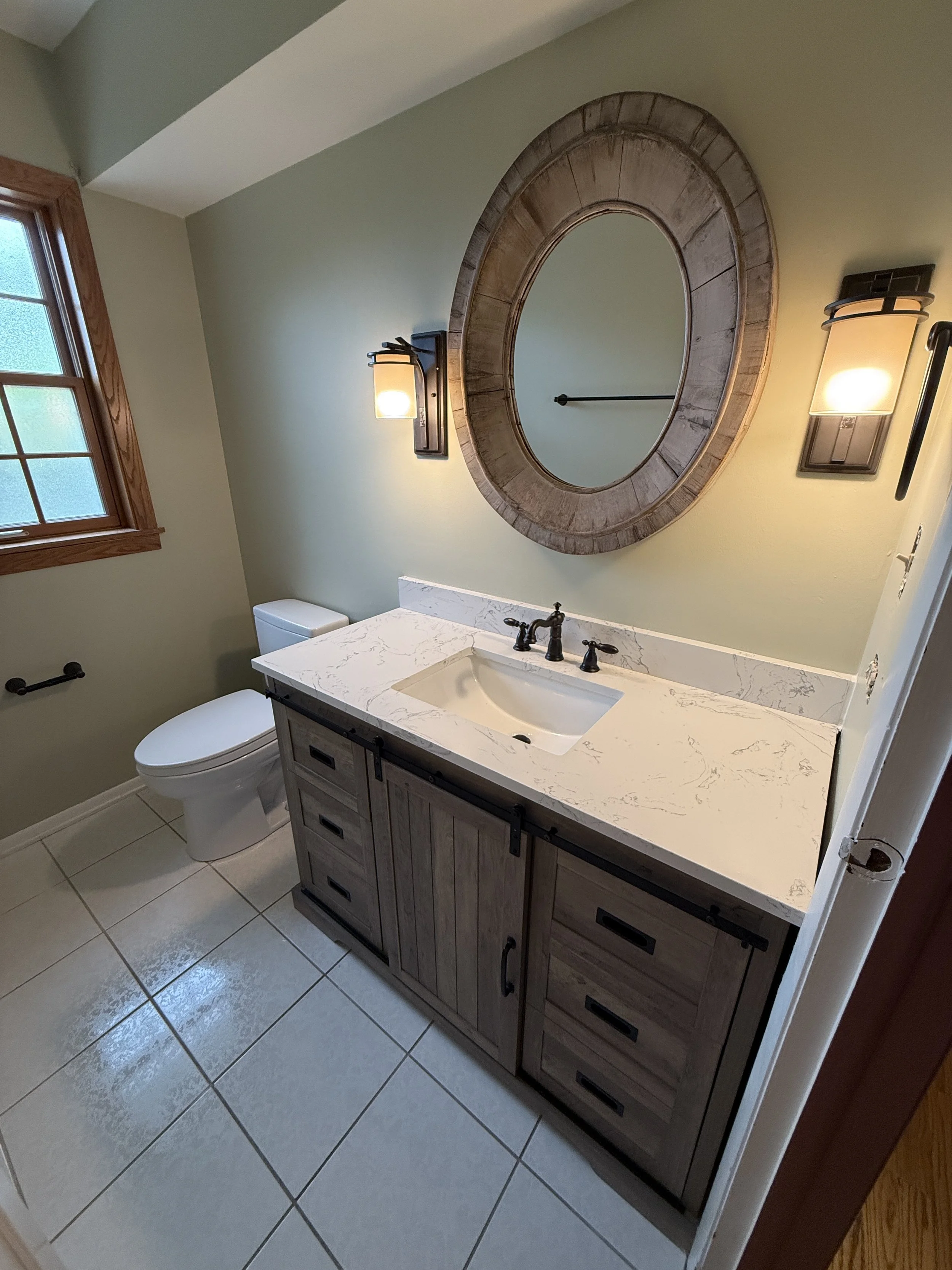 A bathroom with a wooden-framed window, a light green wall, a wooden framed circular mirror above the sink, two wall-mounted light fixtures on either side of the mirror, a white toilet next to the vanity, and a white marble countertop with a built-in sink. The flooring is tiled in light gray.