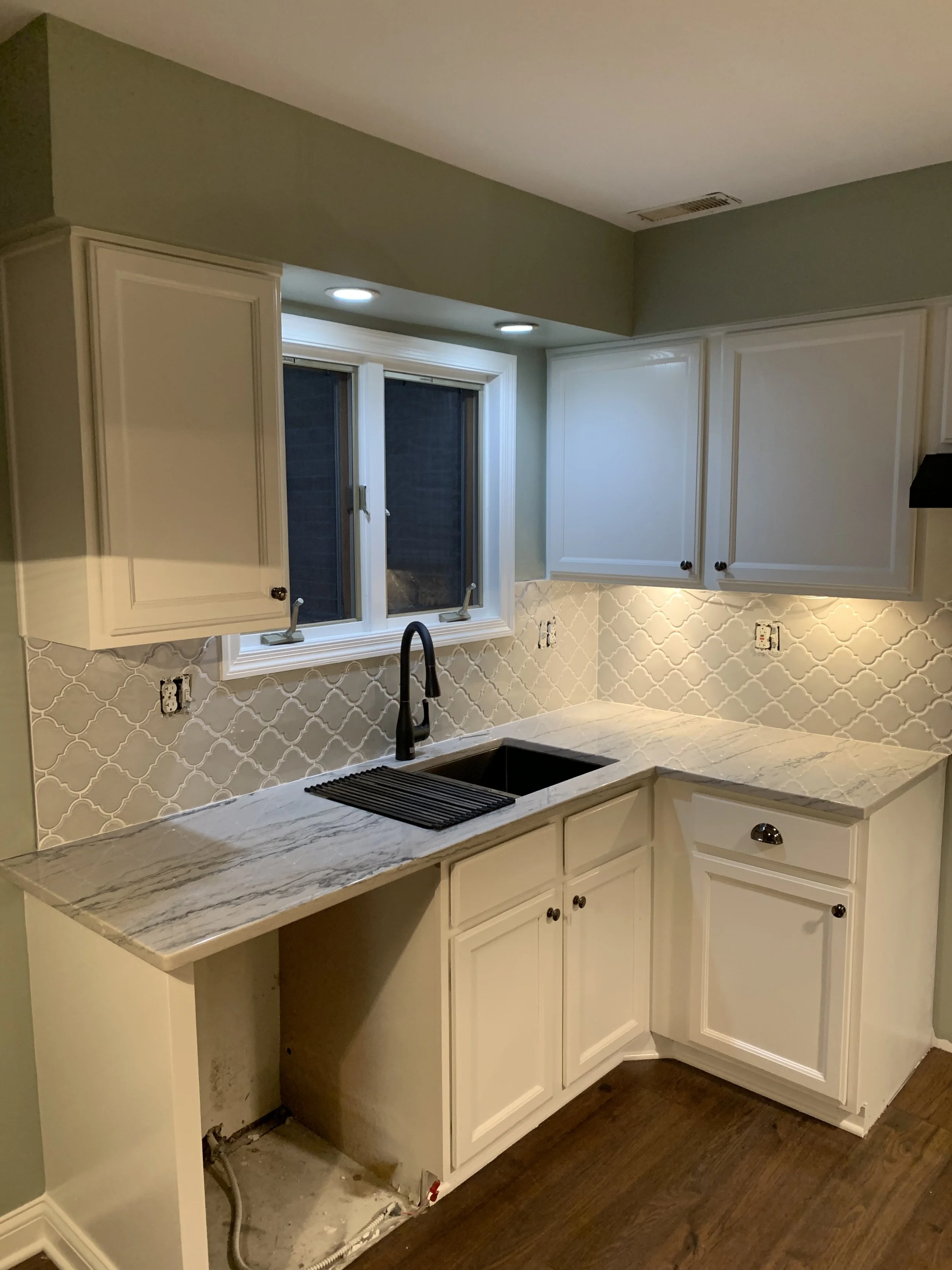 Kitchen with white cabinets, marble countertop, window above sink, black faucet, and beige tile backsplash; under-cabinet lights and dark wood flooring.