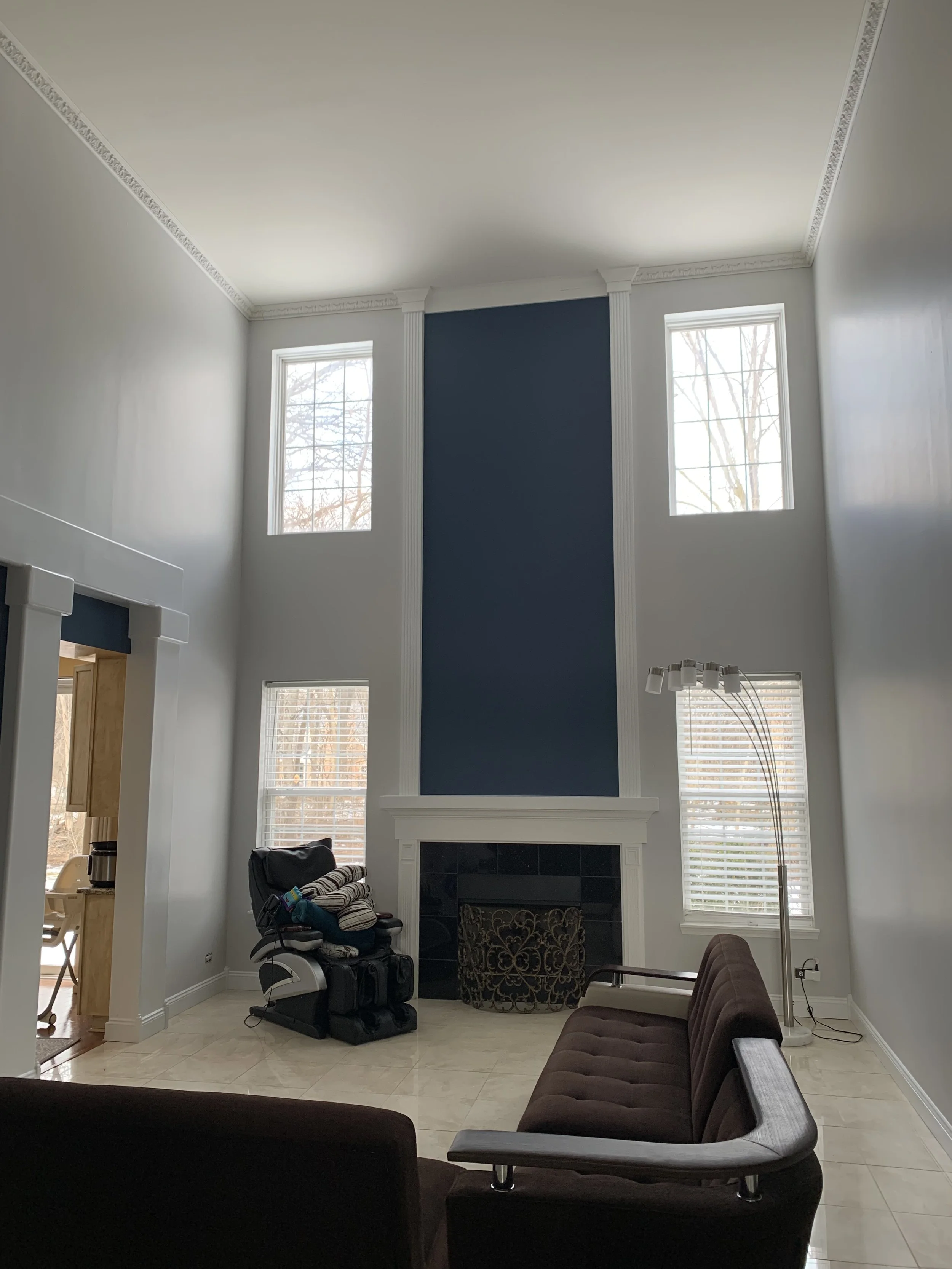 Living room with high ceiling, large windows, a fireplace with a blue accent wall, brown seating, a massage chair, and a floor lamp.