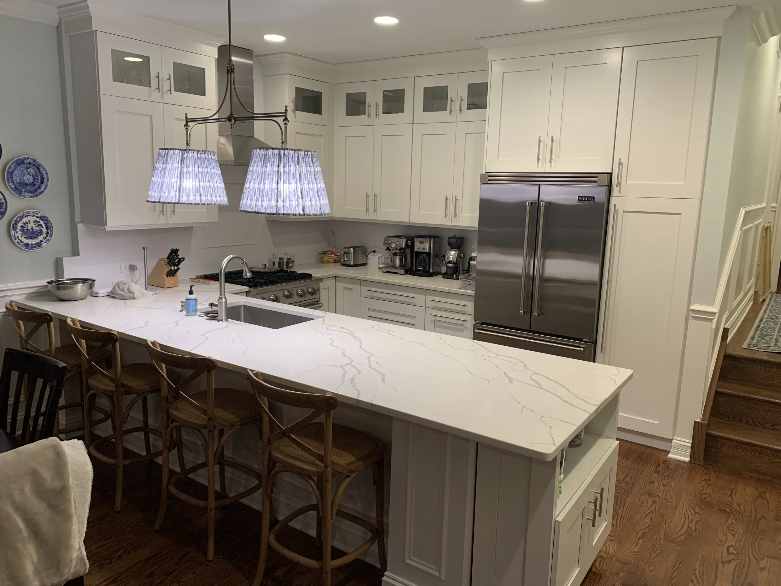 Modern kitchen with white cabinets, marble countertop, stainless steel refrigerator, and wooden bar stools. Two pendant lights hang over the island.