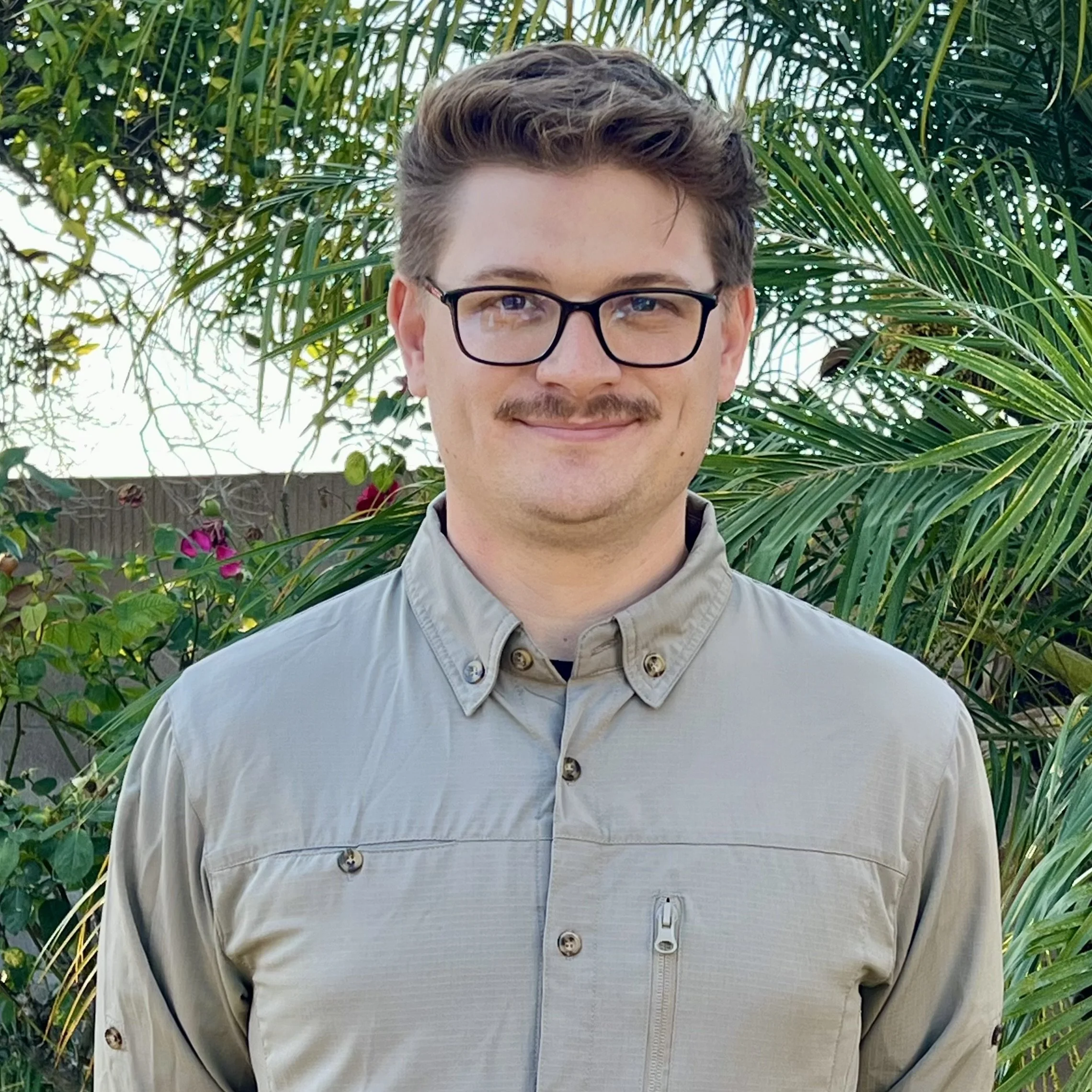 A young man with glasses and a mustache smiling outdoors, wearing a beige button-up shirt with a zipper pocket, standing in front of green tropical plants.