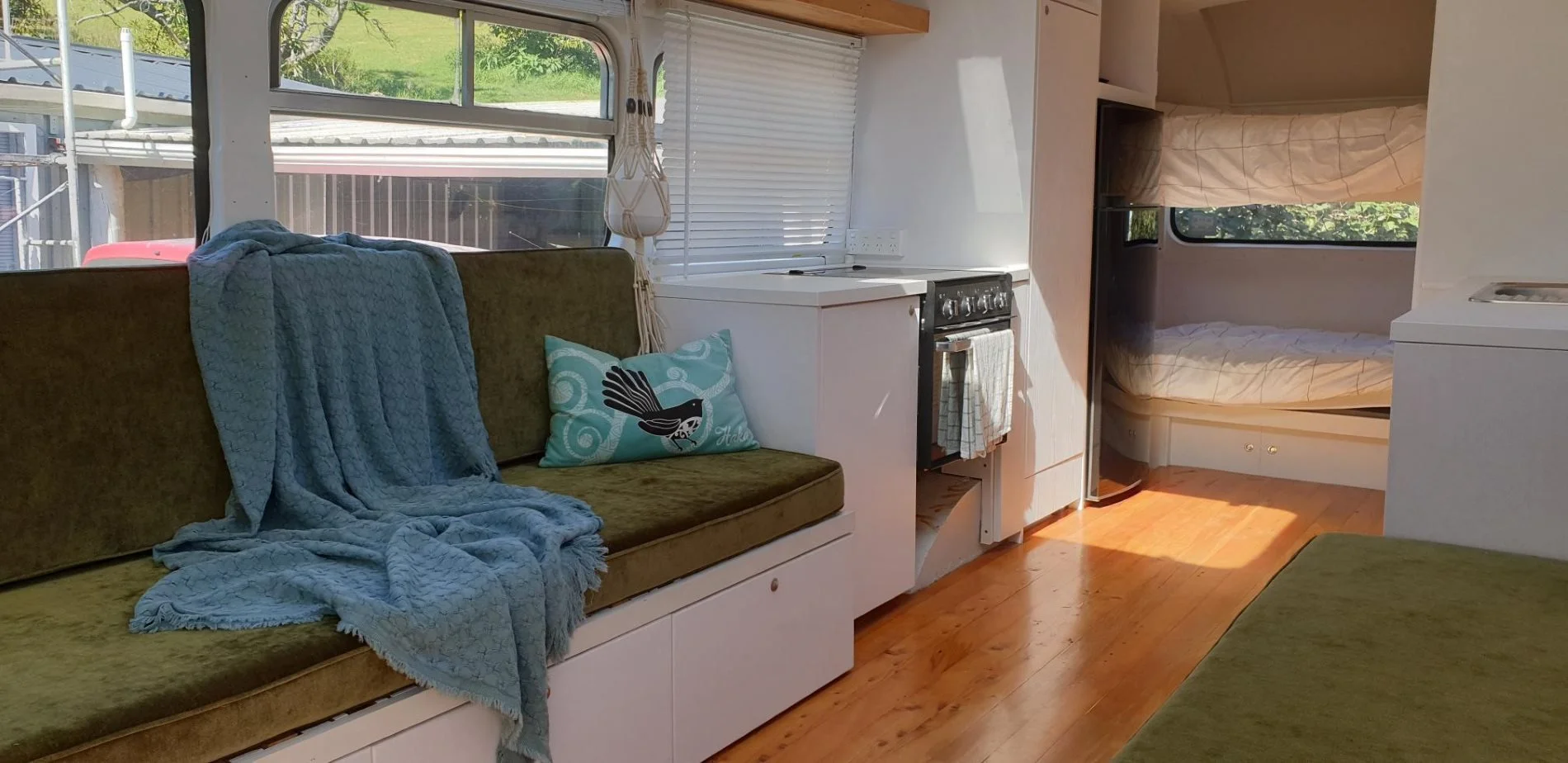 Interior of a small camper with green cushioned seating, a blue blanket, decorative pillow, small kitchen area, and bunk beds at the back