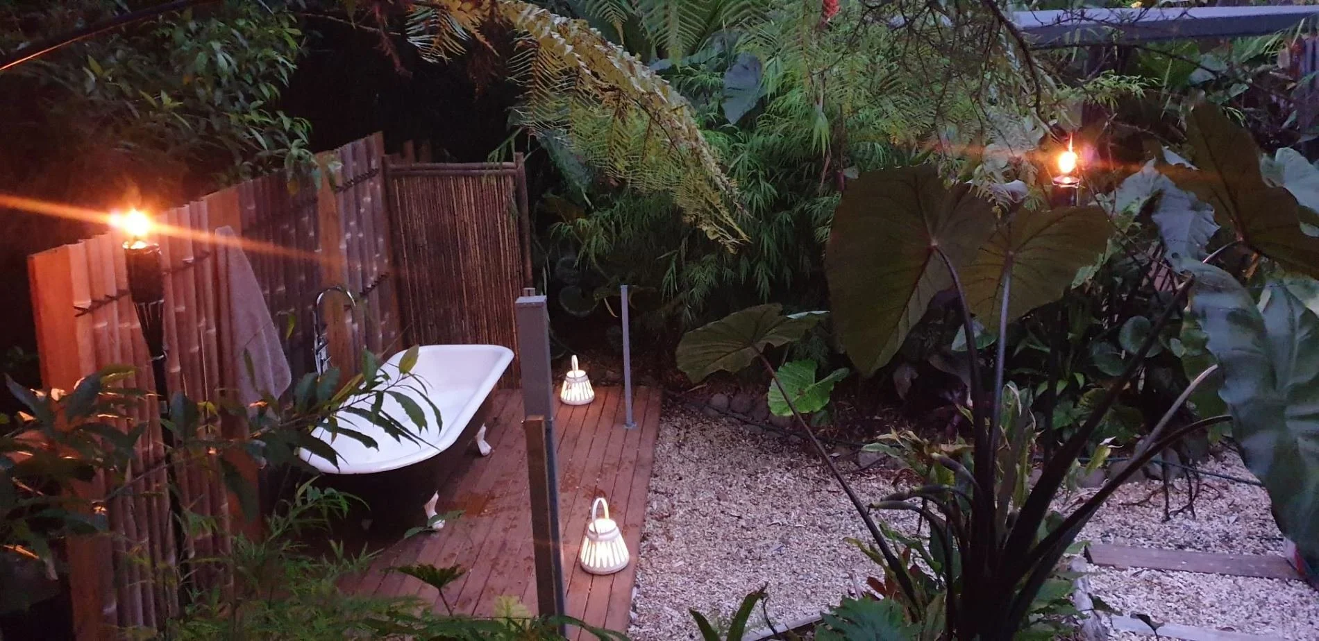 An outdoor garden scene at dusk with a bathtub on a wooden deck, surrounded by lush greenery, tiki torches providing ambient light, and small lanterns on the ground, creating a cozy and tropical atmosphere.