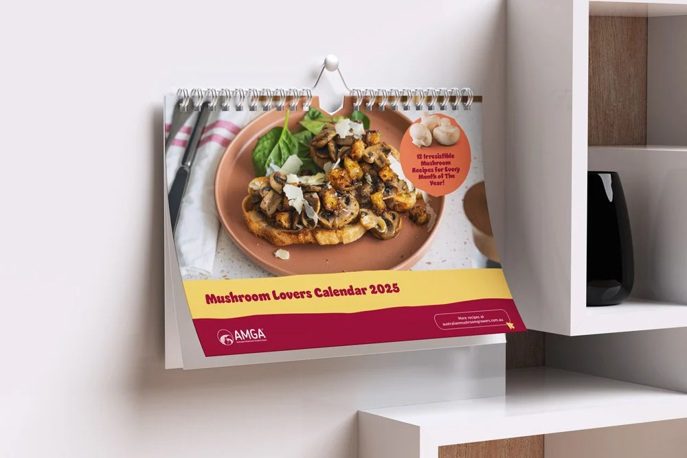 A desk calendar titled "Mushroom Lovers Calendar 2023" showing a plate of cooked mushrooms garnished with cheese, on a pink plate with a basil leaf, with a fork and napkin nearby.