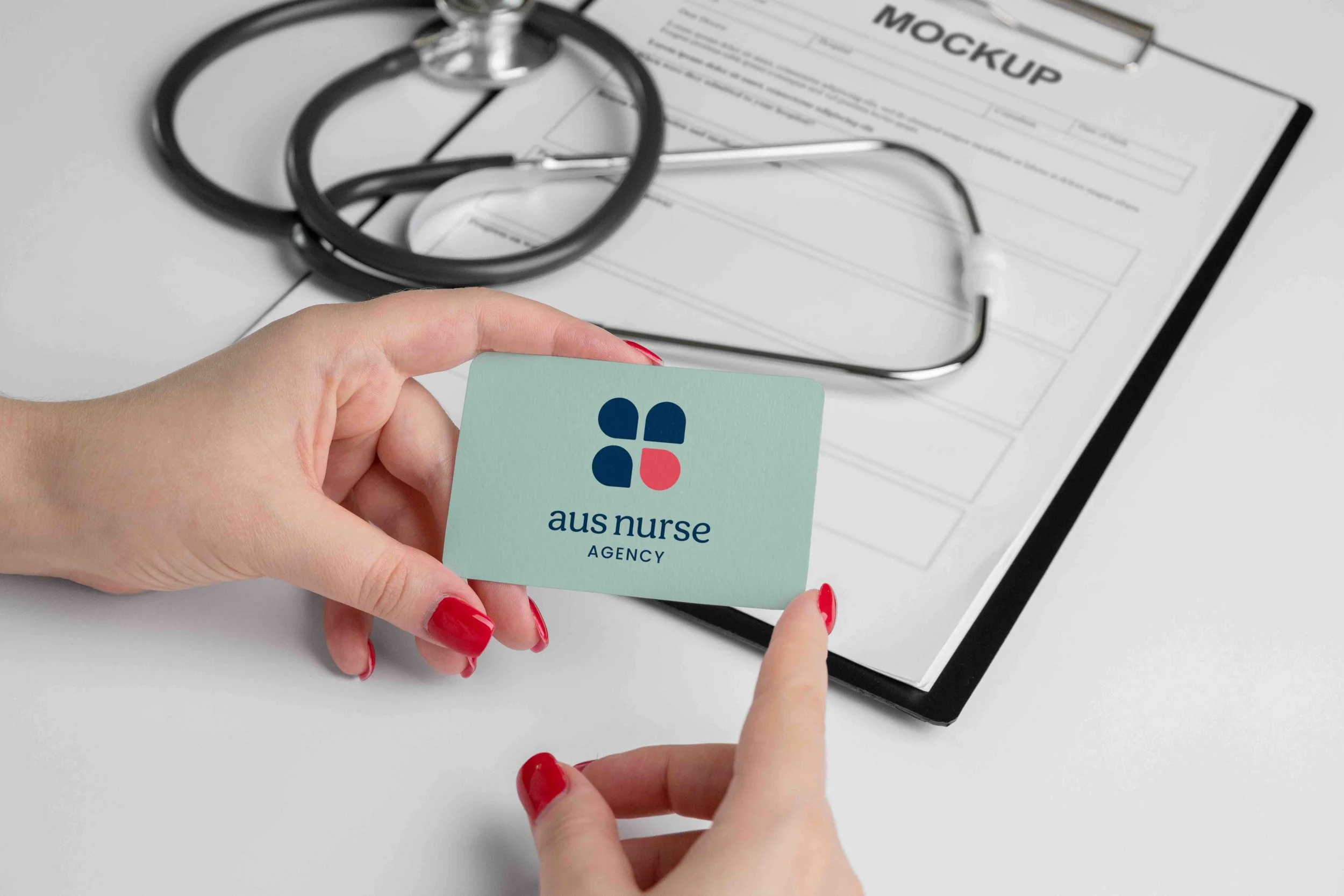 Freelance Graphic Designer Melbourne. Hand holding a business card with 'aus nurse AGENCY' logo, stethoscope and medical document on a white surface.