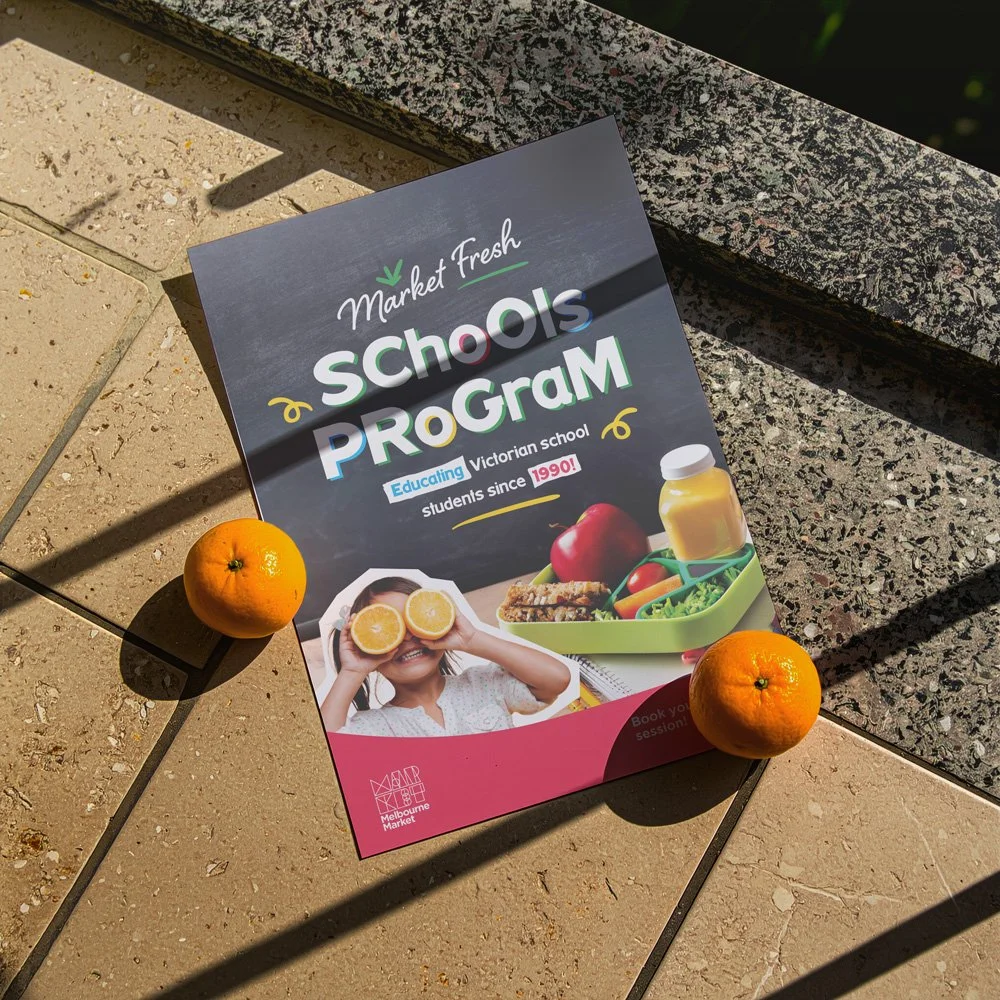 A flyer for a school lunch program placed on a tiled outdoor surface, with two oranges beside it and a granite ledge behind. The flyer promotes a Victorian school lunch program, highlighting its history since 1990, and features images of food and a s