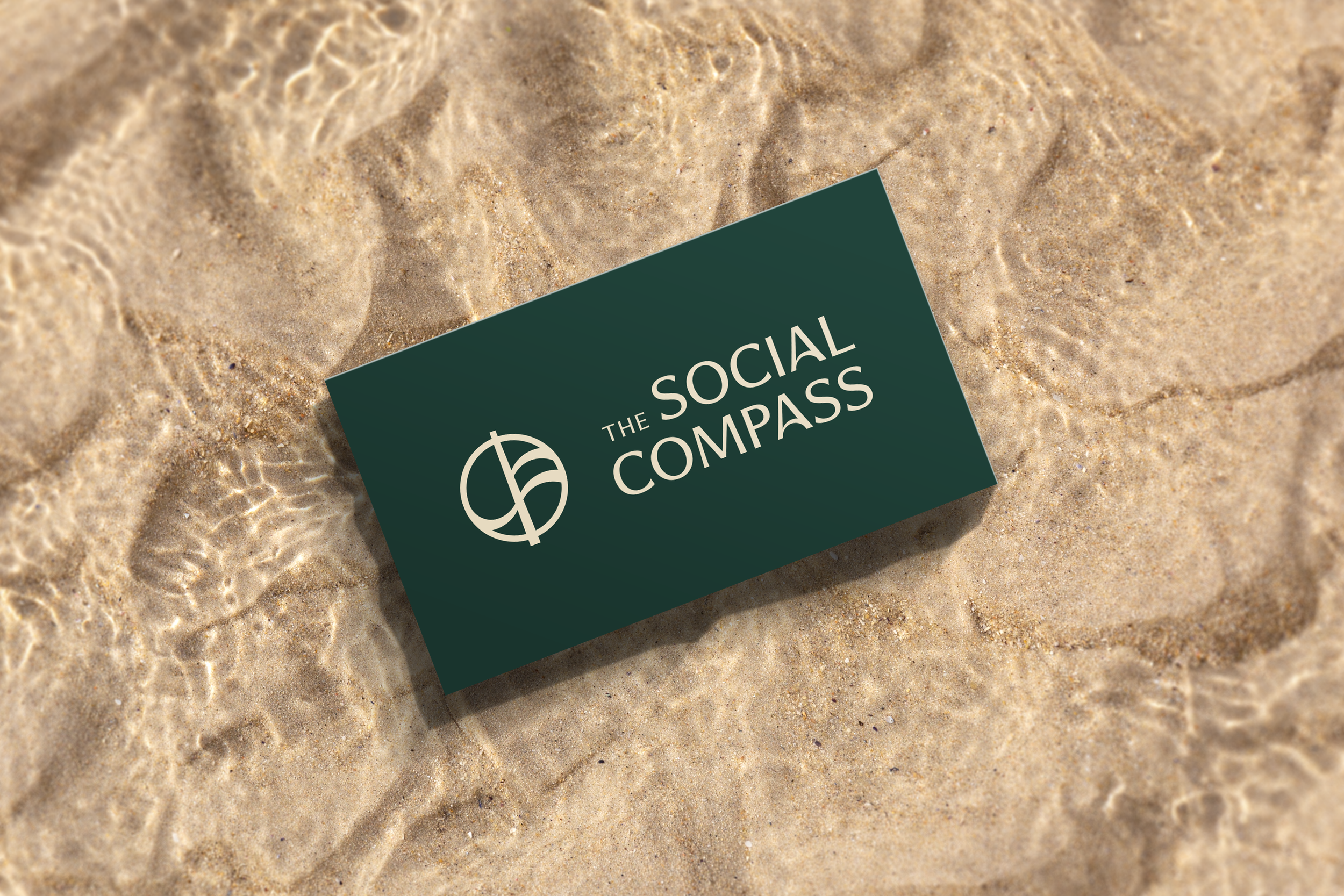 A business card for 'The Social Compass' resting on sandy beach with water ripples. The card features a compass logo on a dark background.