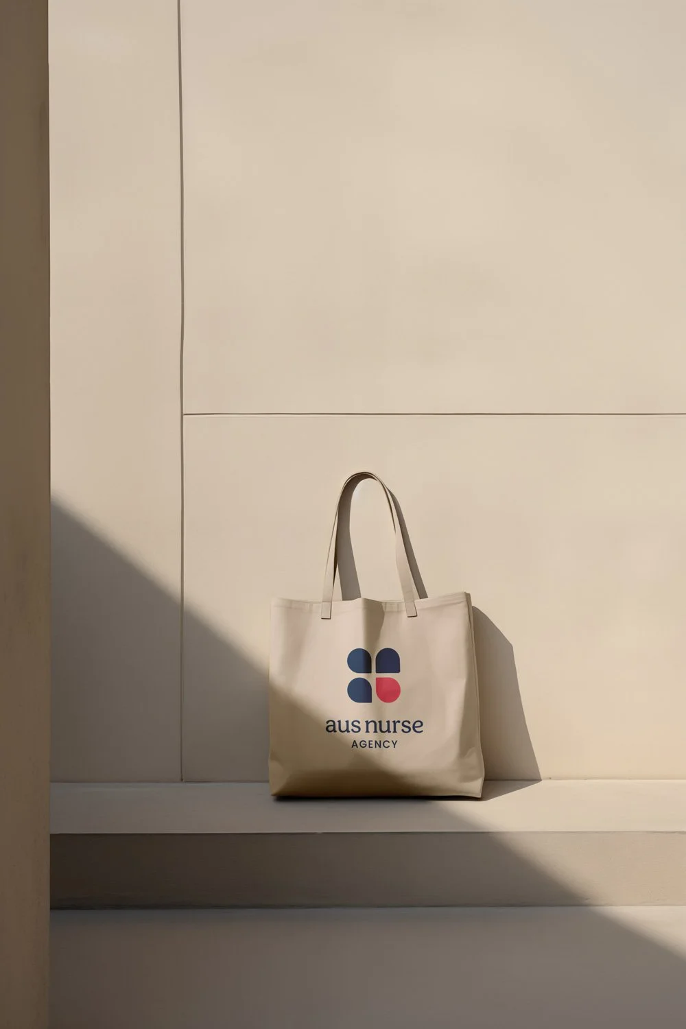 Freelance Graphic Designer Melbourne. A beige tote bag with the logo and text 'aus nurse AGENCY' on it, placed on a ledge against a beige wall, with shadows cast on the wall and bag.