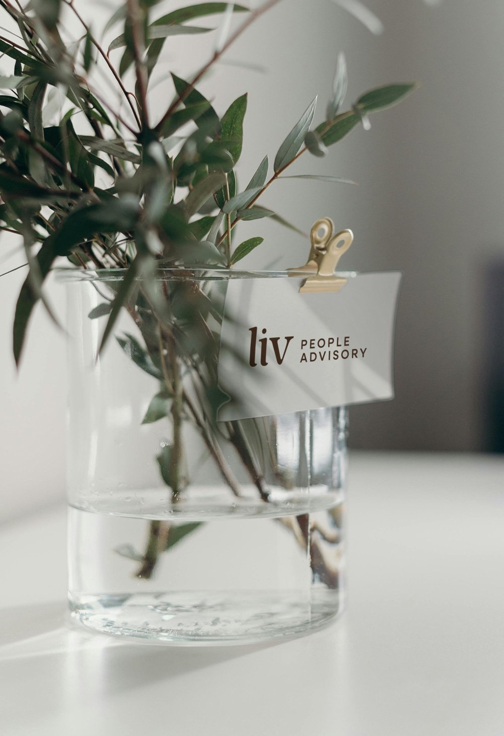 A glass vase with green leafy branches and a small label that reads 'liv PEOPLE ADVISORY' clipped to the vase with a gold binder clip.