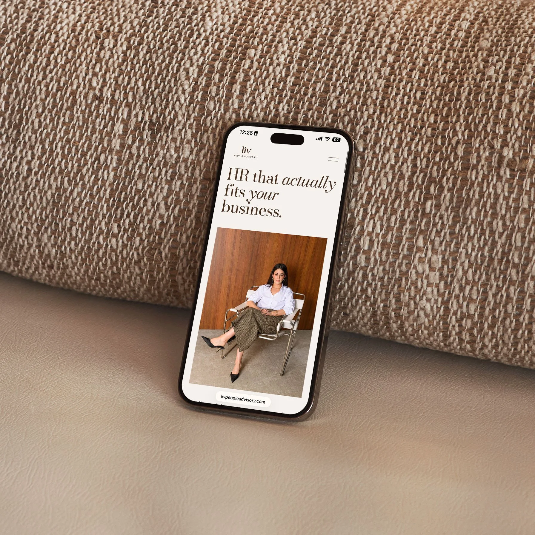 Smartphone displaying a webpage with text about HR fitting your business and a photo of a woman sitting in a chair, on a beige textured surface.