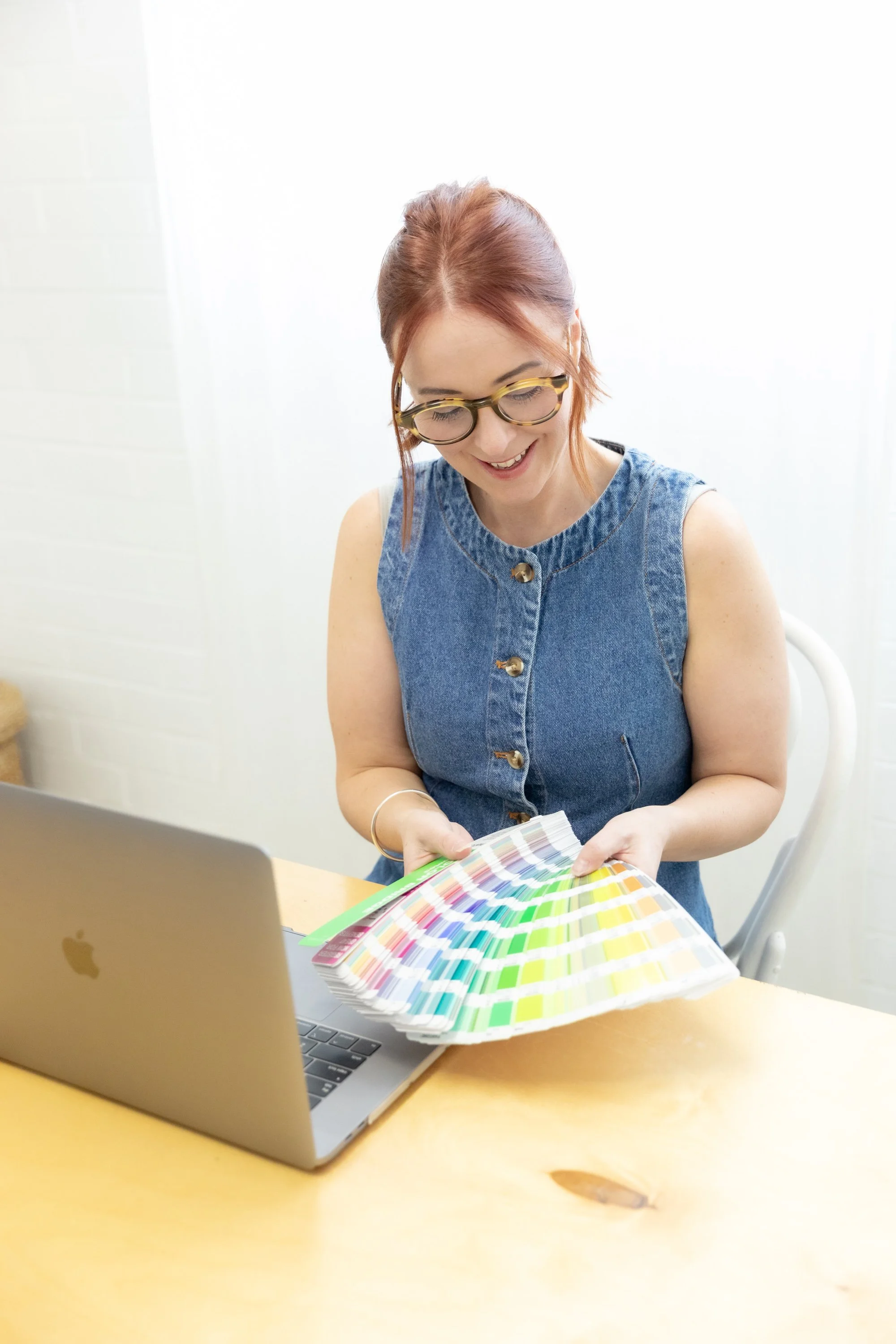 A woman with red hair and glasses looking at a color swatch fan deck while sitting at a wooden table with an open MacBook laptop.