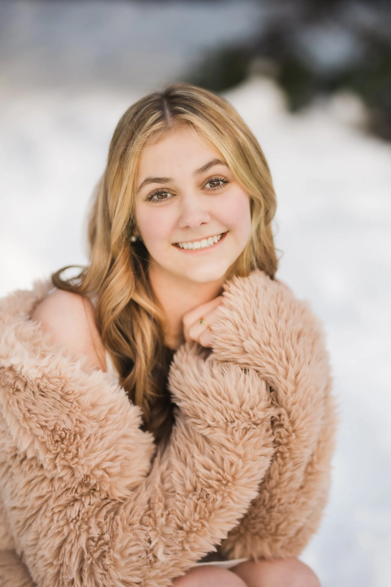 A high school senior girl with long, wavy blonde hair smiling while wearing a cozy jacket outdoors with snow in the background at Gold Creek Pond having her senior photos taken by Cindy Richey Photography